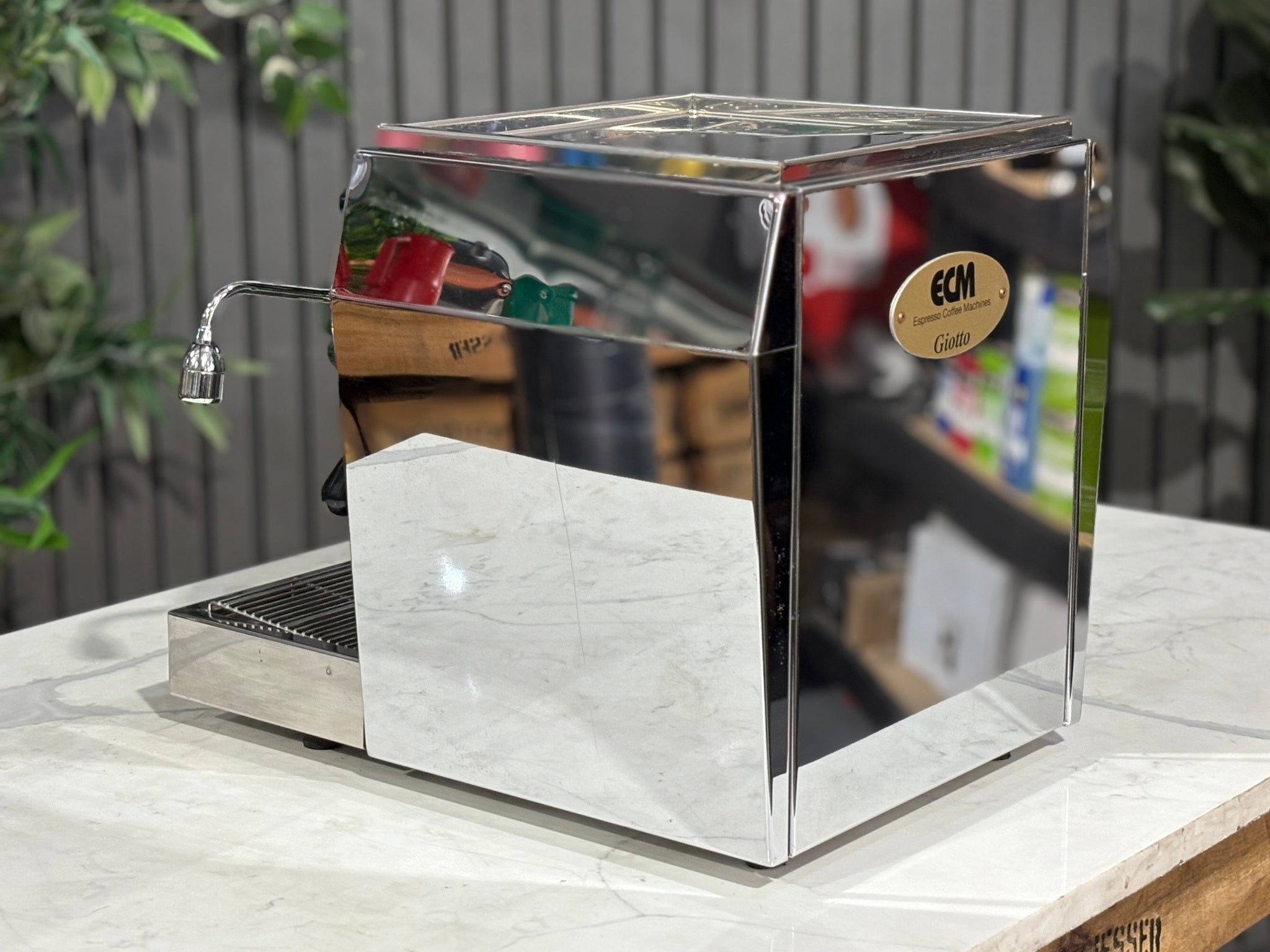 An angled rear image of the ECM Rocket Giotto 1 Group Coffee Machine – Stainless, offering a look at the back and side design features. The stainless body reflects natural light, with the marble surface and plants creating an inviting café-style scene.