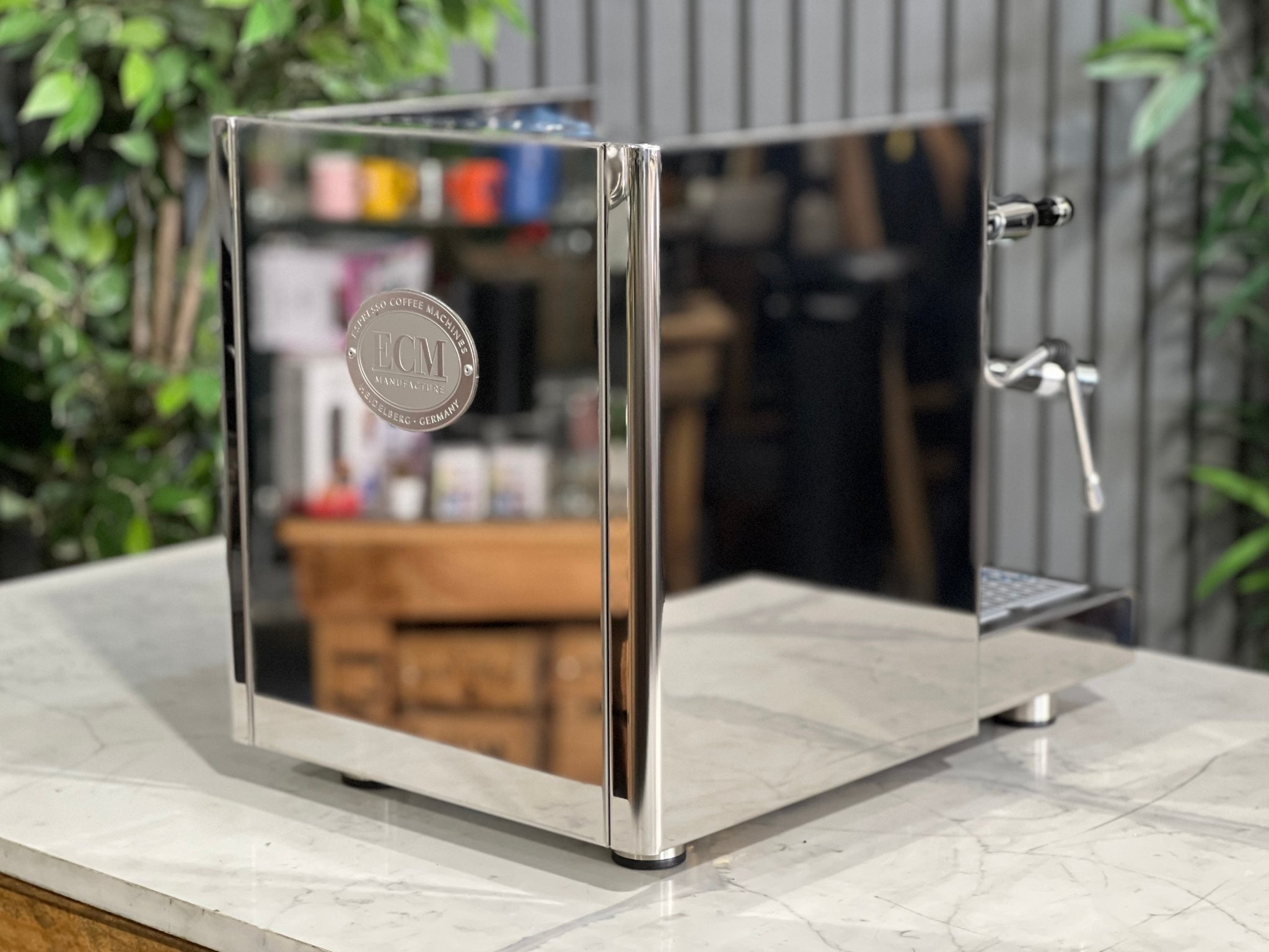 ECM Technika V Profi Rotary PID 1 Group Coffee Machine Stainless – New espresso coffee machine angled from the back, showing the stainless steel rear panel, top cup tray, and part of the E61 group head on a marble bench with greenery behind.