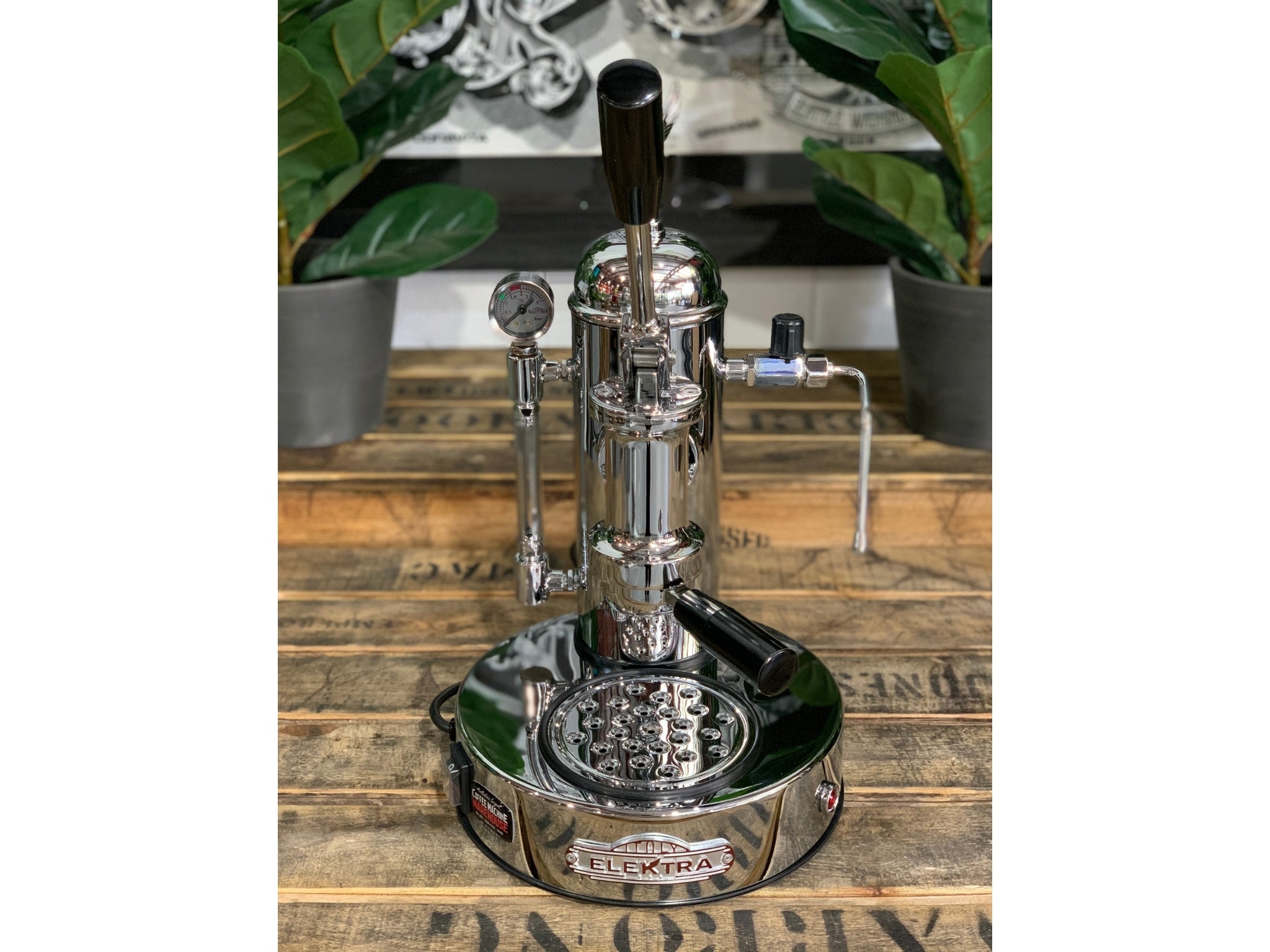 A brand new Elektra Micro Casa Leva 1 Group Coffee Machine with Chrome finish sitting on a wooden bench. There is greenery in the background. The image is taken from the front of the machine so that you can see the front of the machine, and group head.