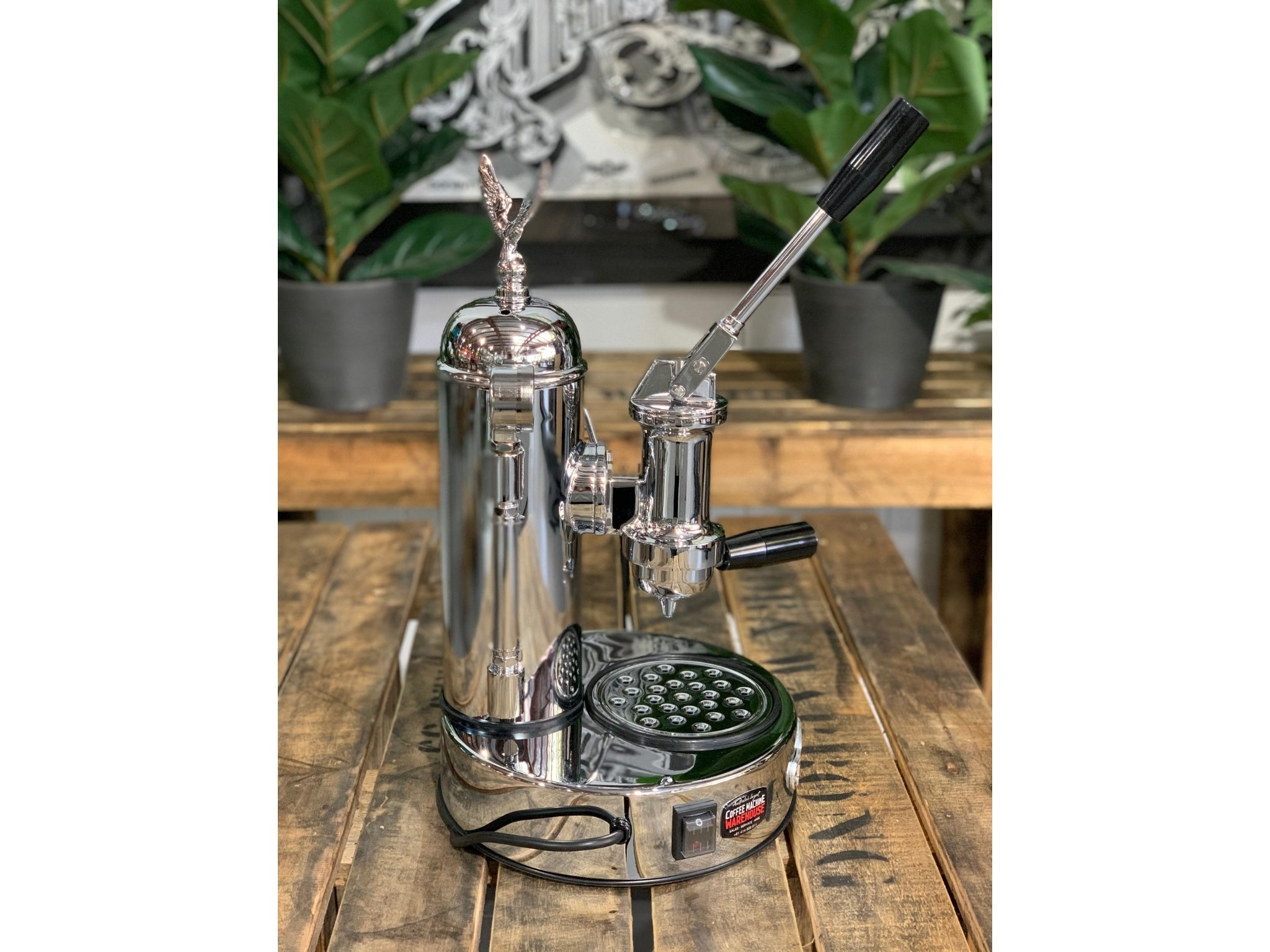A brand new Elektra Micro Casa Leva 1 Group Coffee Machine with Chrome  sitting on a wooden bench. There is greenery in the background. The image is taken from the side of the machine.
