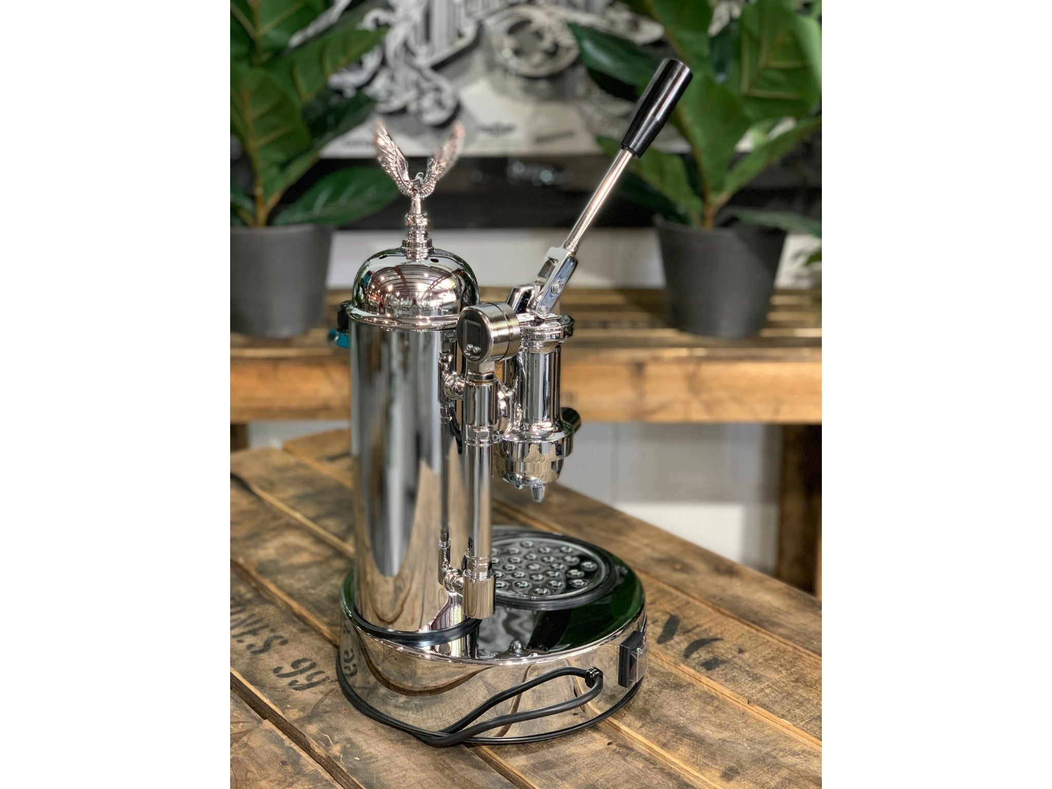 A brand new Elektra Micro Casa Leva 1 Group Coffee Machine with Chrome finish sitting on a wooden bench. There is greenery in the background. The image is taken from behind the machine and the machine is sitting on an angle so that you can see the back of the machine and the side.