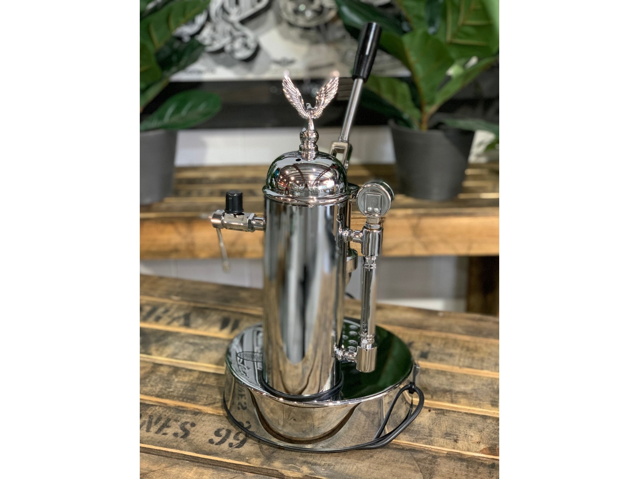 A brand new Elektra Micro Casa Leva 1 Group Coffee Machine with Chrome finishis sitting on a wooden bench. There is greenery in the background. The image is taken from behind the machine so the back is visible.