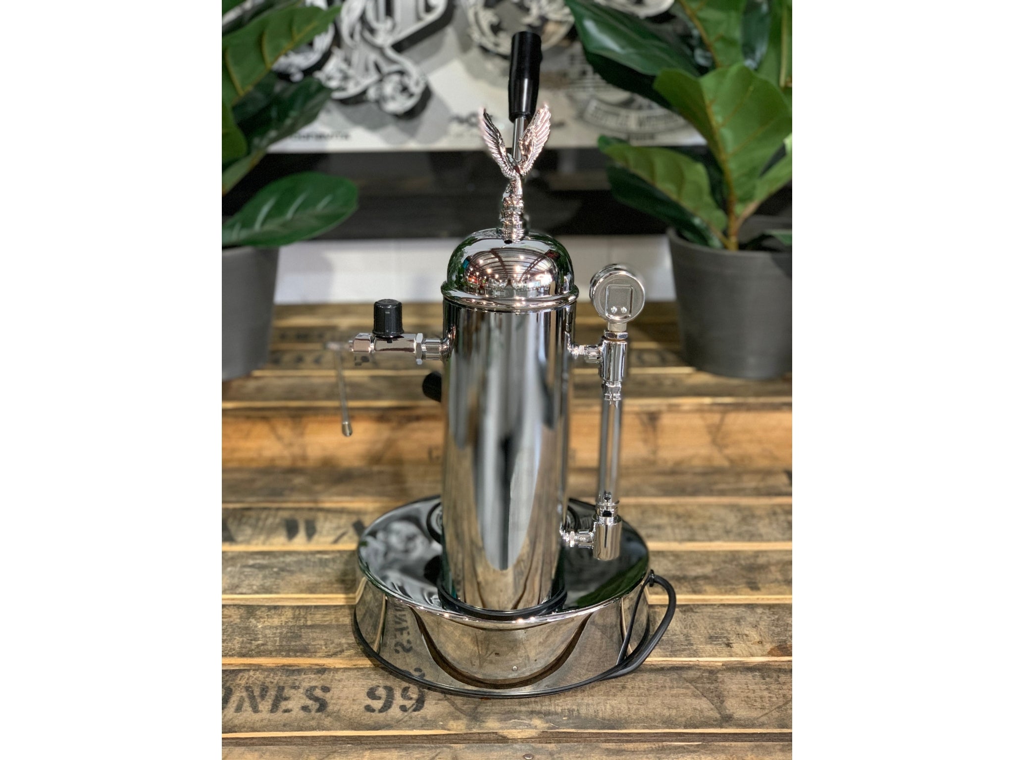 A brand new Elektra Micro Casa Leva 1 Group Coffee Machine with Chrome finish is sitting on a wooden bench. There is greenery in the background. The image is taken from behind the machine so the back is visible
