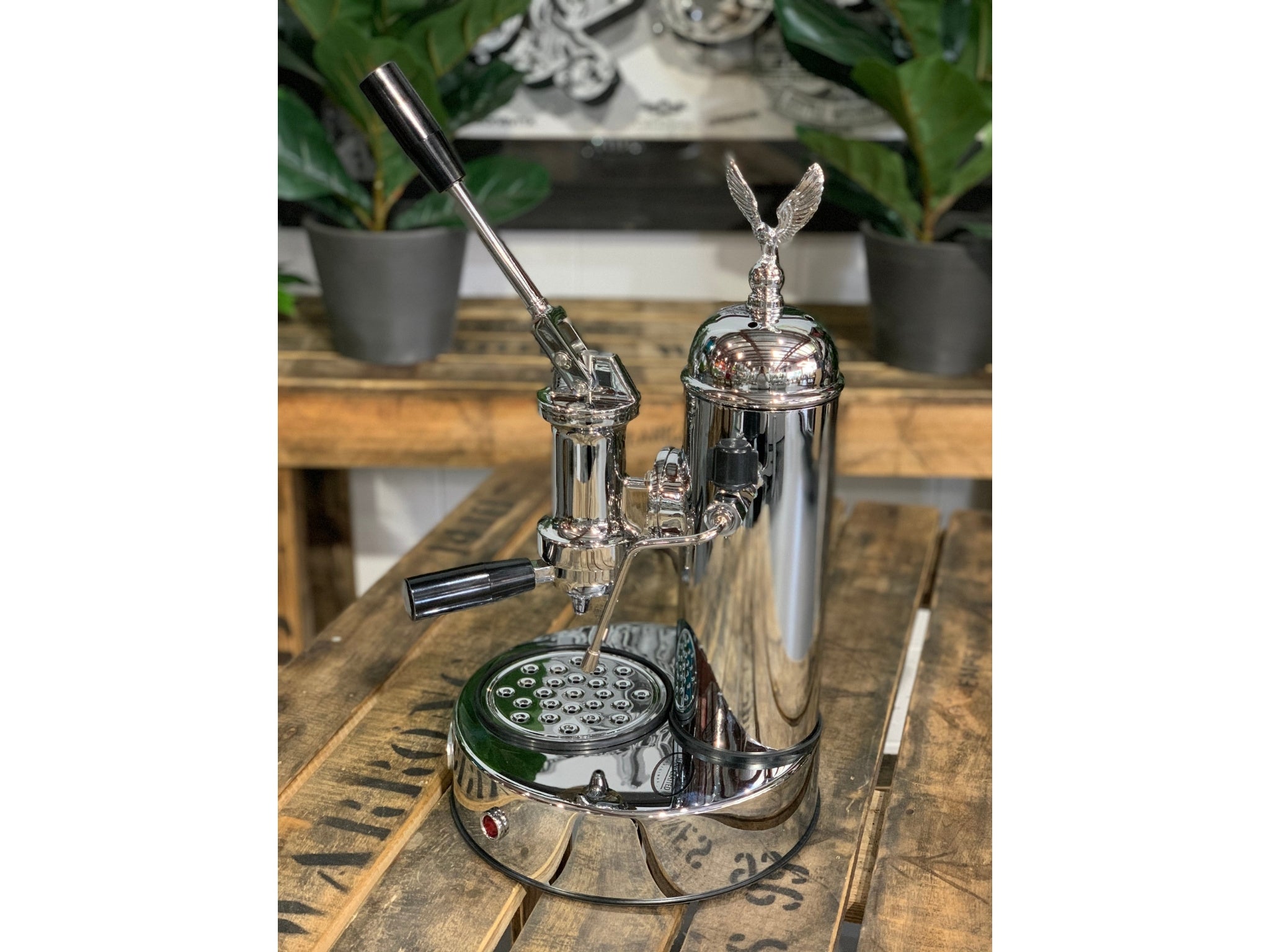 A brand new Elektra Micro Casa Leva 1 Group Coffee Machine with Chrome finish sitting on a wooden bench. There is greenery in the background. The image is taken from the side of the machine.
