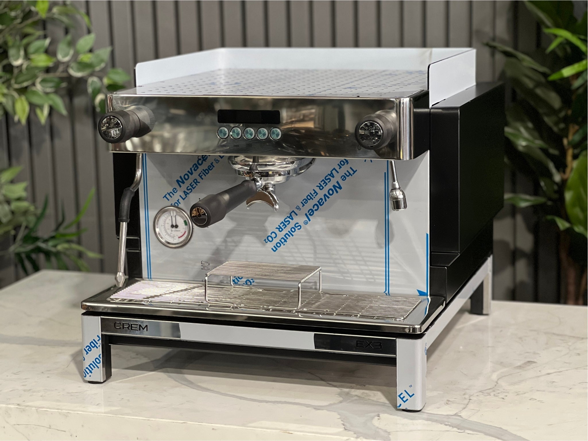 Expobar Crem EX3 Compact 1 Group Coffee Machine Black - New angled view showing the sleek black body, high feet, steam wand, and control panel on a marble bench with greenery in the background. A compact commercial espresso coffee machine built for professional use.