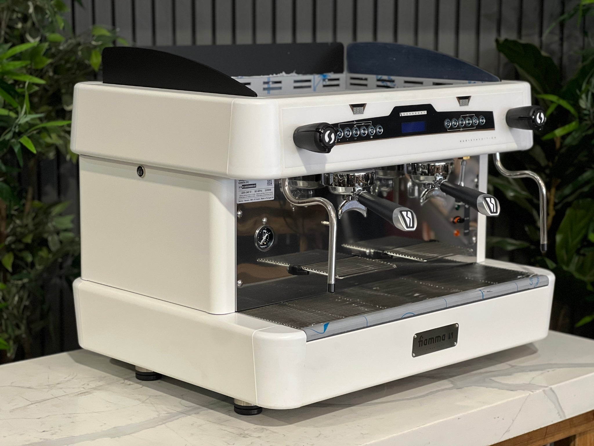 A brand new Fiamma Quadrant 2 DSP 2 Group Coffee Machine with White finish
is sitting on a marble bench. There is greenery in the background. The image is taken from the front of the machine and the machine is sitting on an angle so that you can see the front of the machine and the side. 