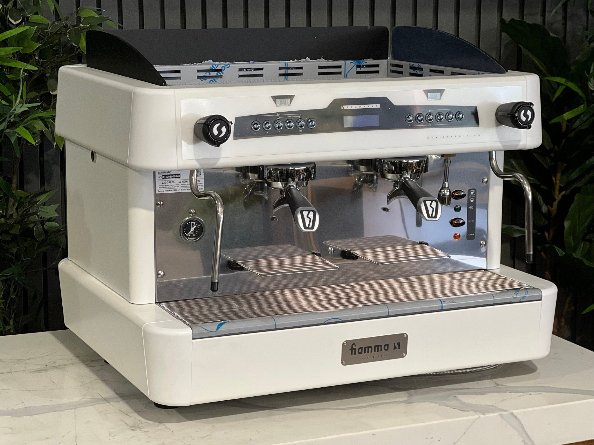 A brand new Fiamma Quadrant 2 DSP 2 Group Coffee Machine with White finish
is sitting on a marble bench. There is greenery in the background. The image is taken from the front of the machine and the machine is sitting on an angle so that you can see the front of the machine and the side. 