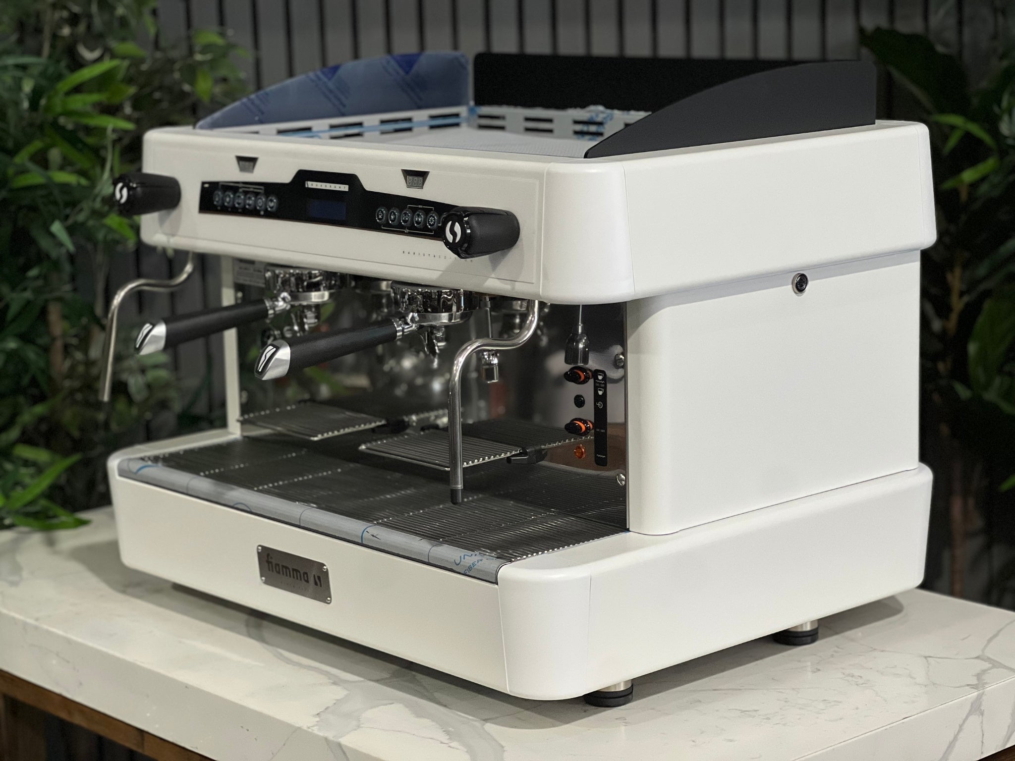 A brand new Fiamma Quadrant 2 DSP 2 Group Coffee Machine with White finish
is sitting on a marble bench. There is greenery in the background. The image is taken from the front of the machine and the machine is sitting on an angle so that you can see the front of the machine and the side. 
