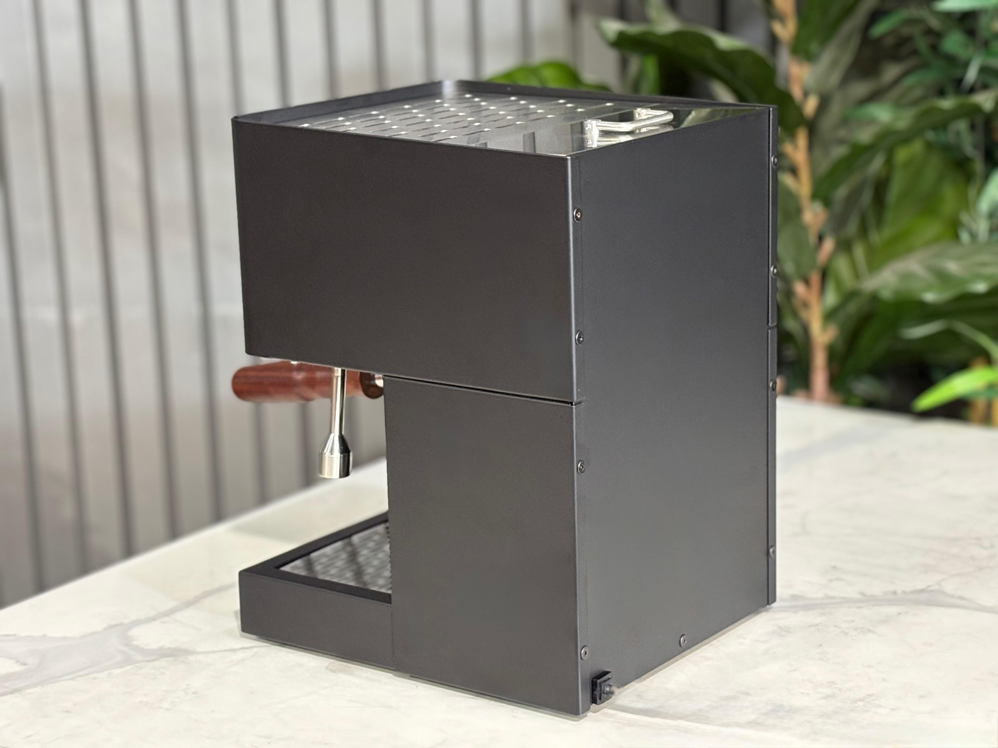 Essenza Nina 1 Group Coffee Machine Stainless side view on a countertop with greenery in the background.