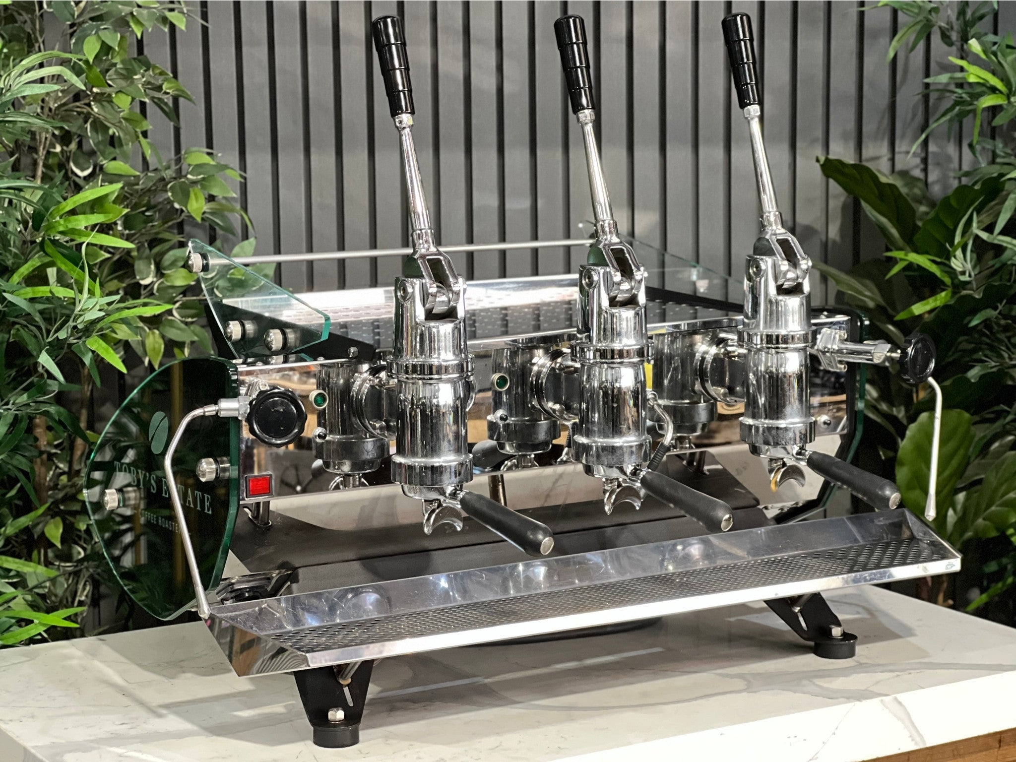Kees van der Westen Mirage Idrocompresso Triplette 3 Group Coffee Machine Stainless - Used espresso machine shown at an angled view on a marble bench, with greenery in the background. Visible features include the stainless steel body, three group heads, dual steam wands, and spring lever mechanisms.