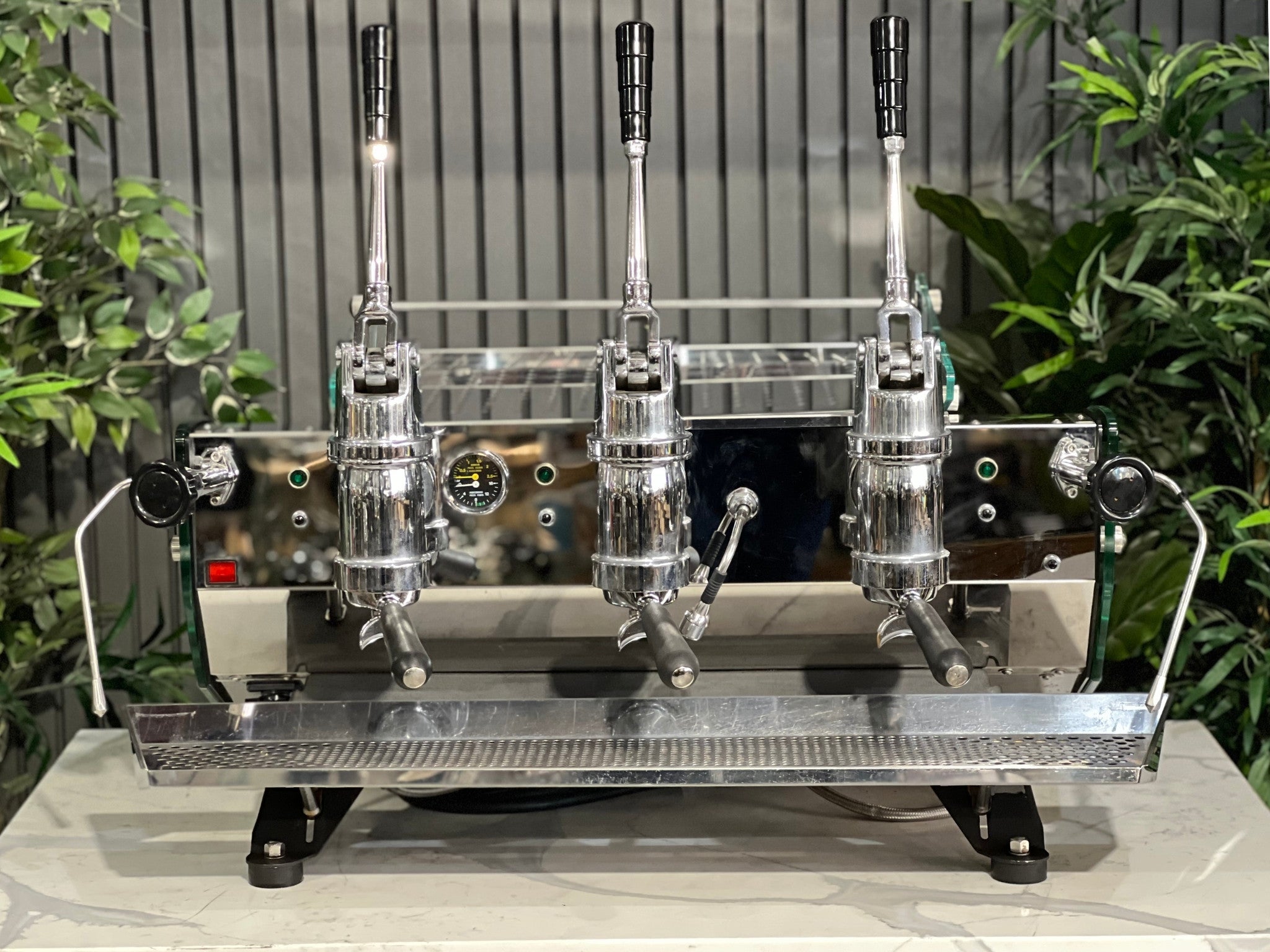 Kees van der Westen Mirage Idrocompresso Triplette 3 Group Coffee Machine Stainless - Used espresso machine shown at an angled view on a marble bench, with greenery in the background. Visible features include the stainless steel body, three group heads, dual steam wands, and spring lever mechanisms.
