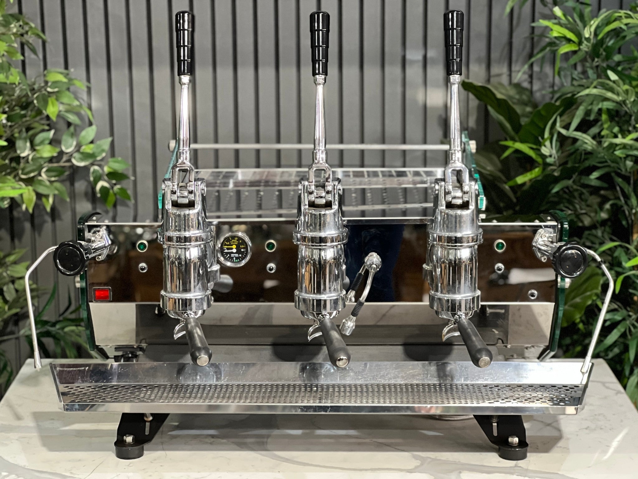 Kees van der Westen Mirage Idrocompresso Triplette 3 Group Coffee Machine Stainless - Used espresso machine shown from a slightly elevated front view. Highlights the top panel, lever assemblies, and cup tray surface, set against a marble bench with greenery behind.