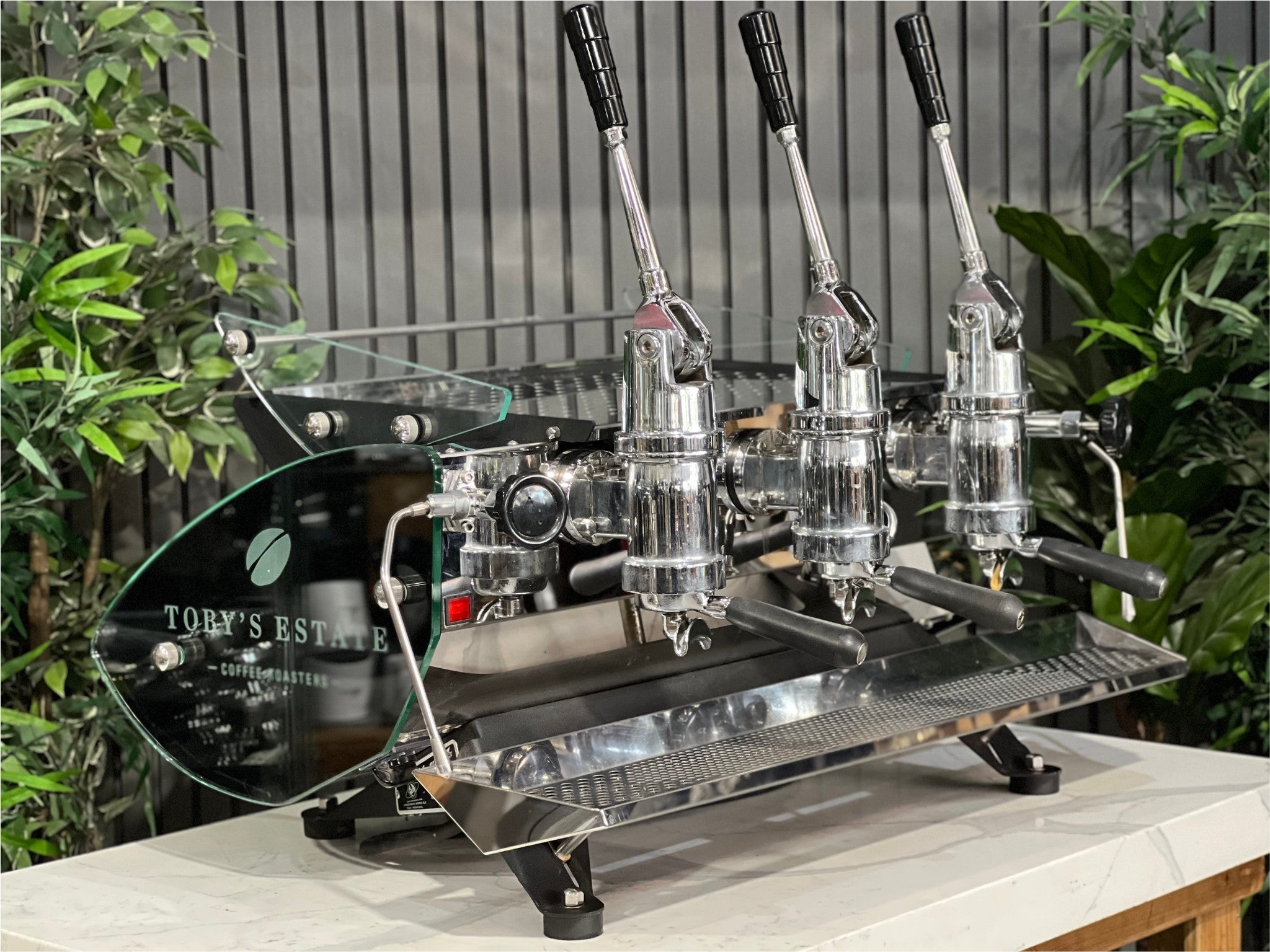 Kees van der Westen Mirage Idrocompresso Triplette 3 Group Coffee Machine Stainless - Used espresso machine shown at a front-left angle on a marble bench, with greenery in the background. The stainless steel body, lever handles, triple group heads, and dual steam wands are clearly visible.