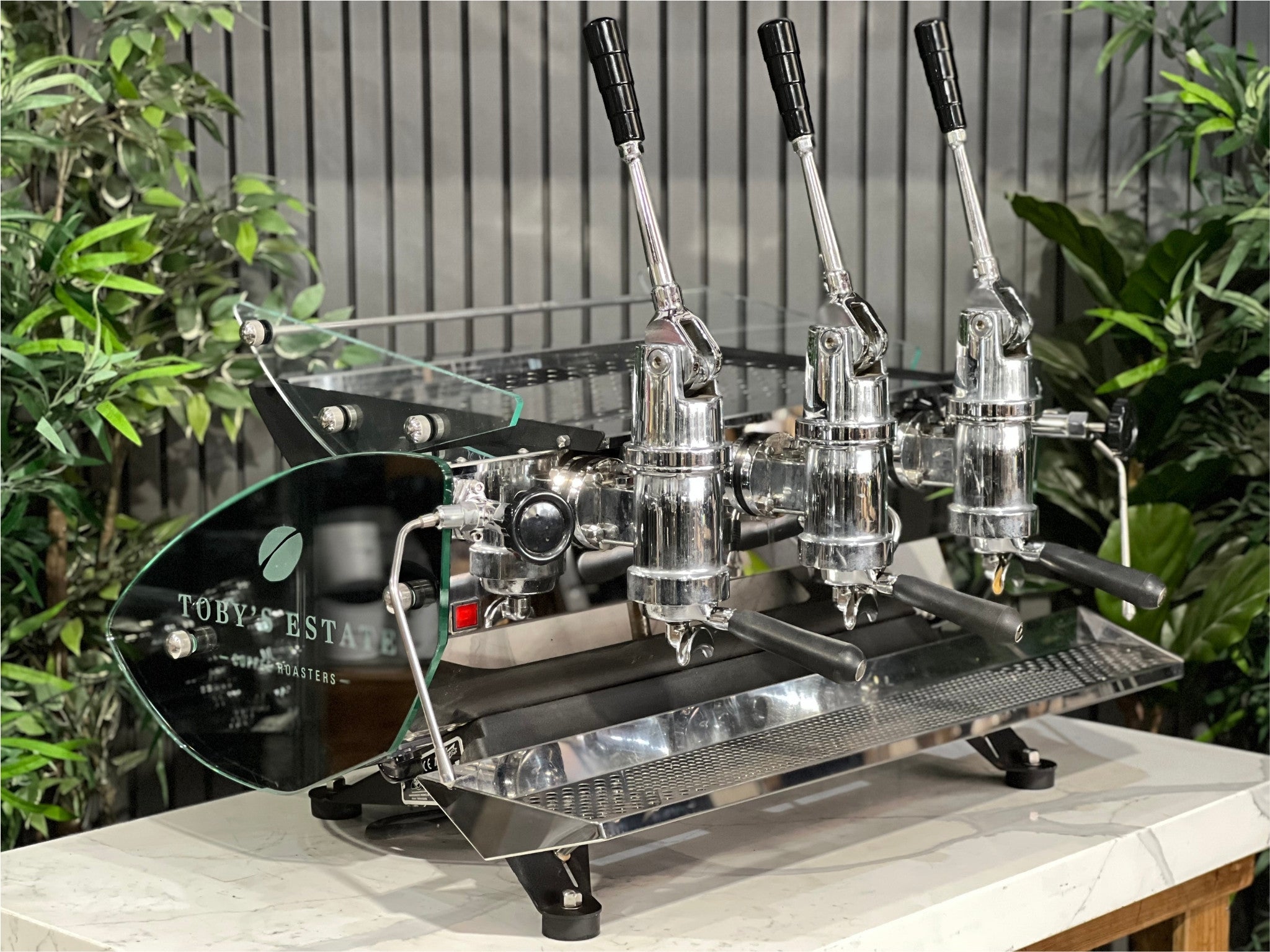 Kees van der Westen Mirage Idrocompresso Triplette 3 Group Coffee Machine Stainless - Used espresso machine shown at a front-left angle on a marble bench, with greenery in the background. The stainless steel body, lever handles, triple group heads, and dual steam wands are clearly visible.