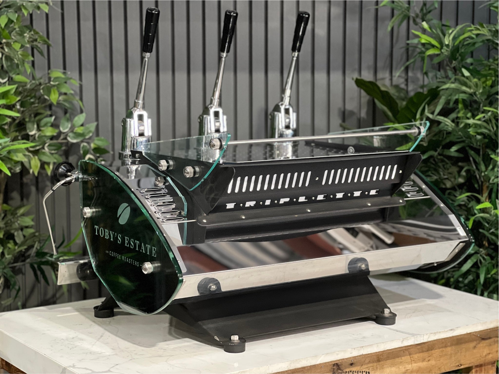 Kees van der Westen Mirage Idrocompresso Triplette 3 Group Coffee Machine Stainless - Used espresso machine photographed from an angled back perspective. Emphasises the contours of the stainless body, rear structure, and lever design, with a marble bench and greenery behind.