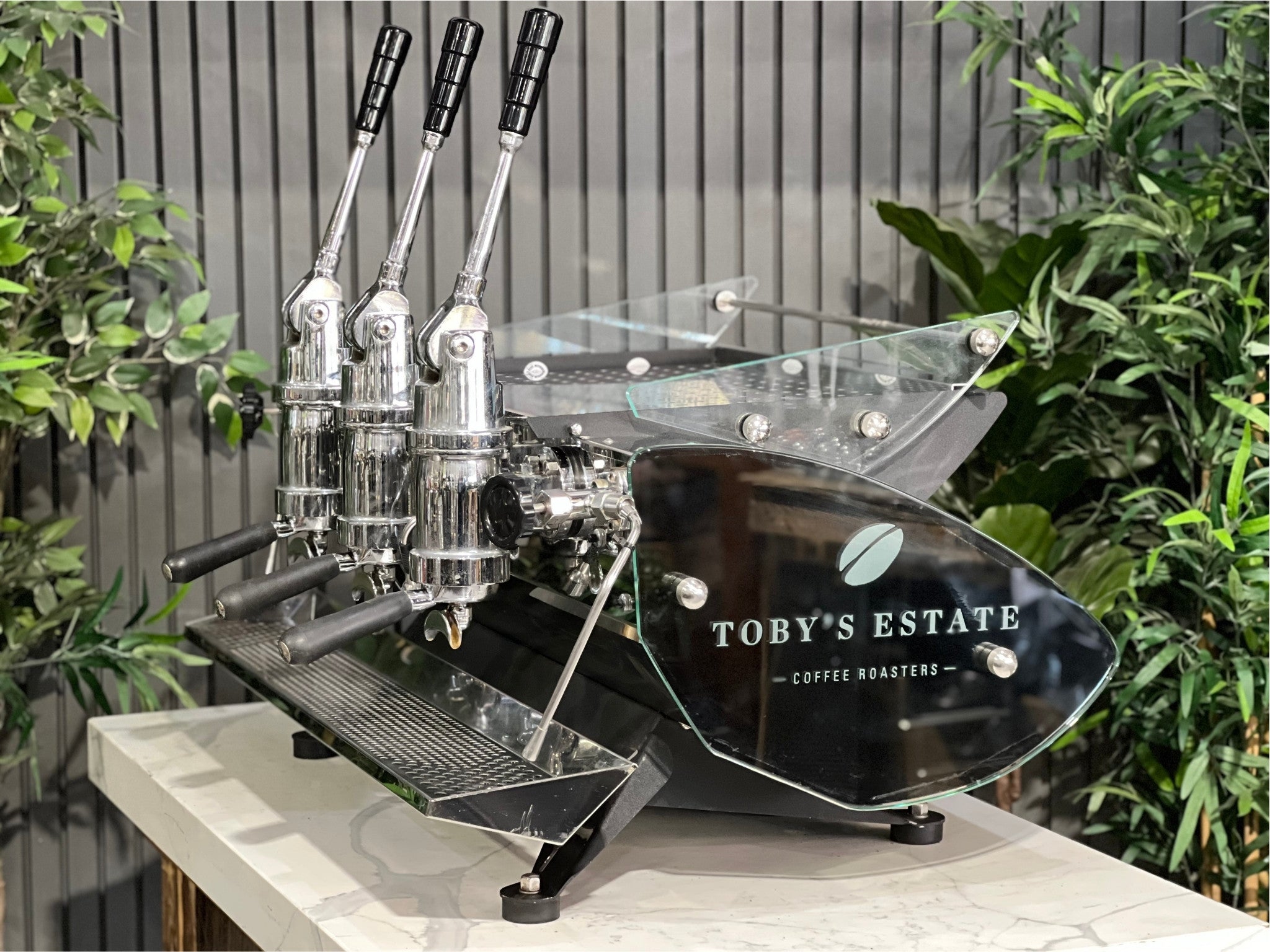 Kees van der Westen Mirage Idrocompresso Triplette 3 Group Coffee Machine Stainless - Used espresso machine shown at an angled view on a marble bench, with greenery in the background. Visible features include the stainless steel body, three group heads, dual steam wands, and spring lever mechanisms.
