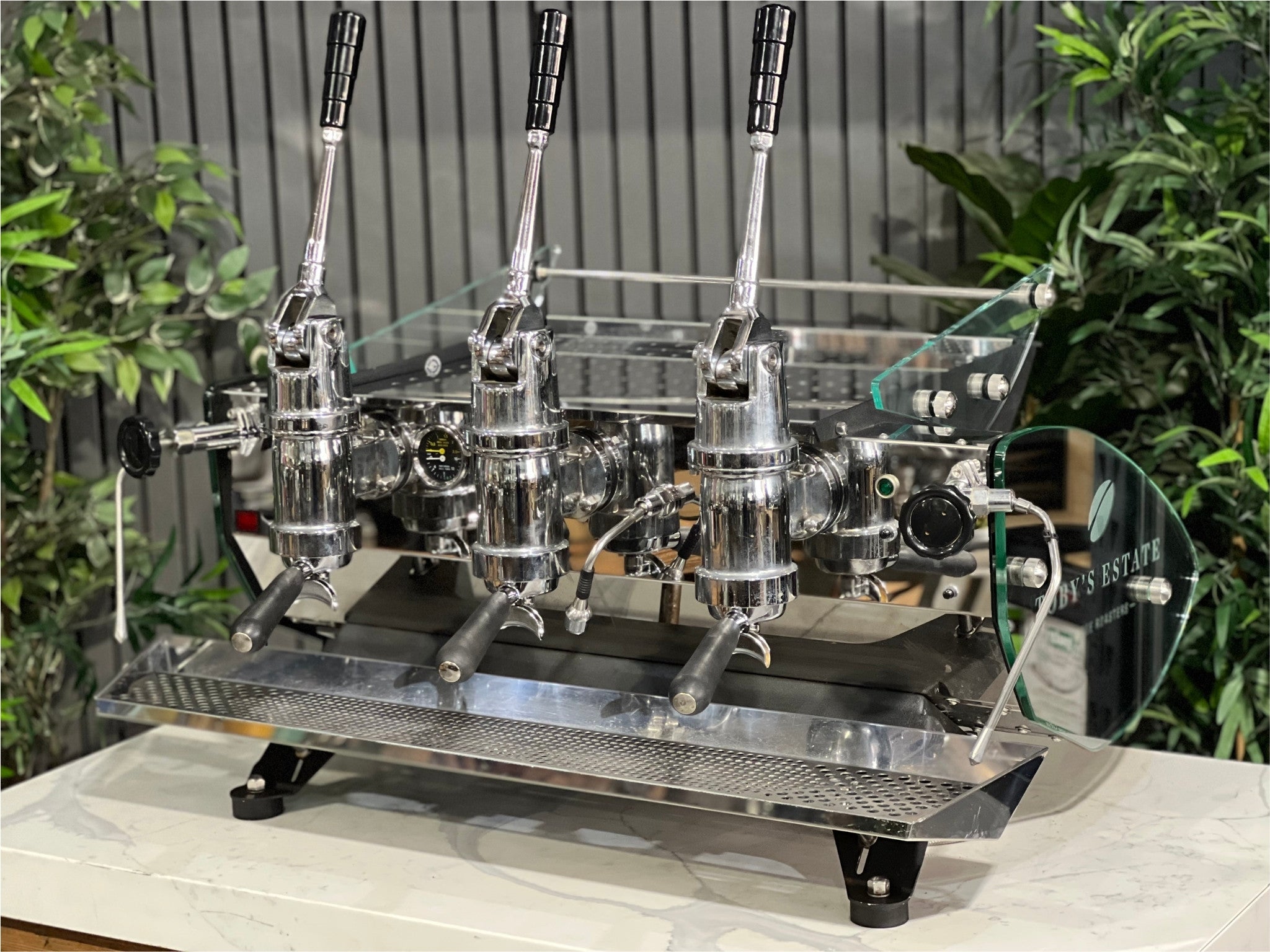 Kees van der Westen Mirage Idrocompresso Triplette 3 Group Coffee Machine Stainless - Used espresso machine shown at an angled view on a marble bench, with greenery in the background. Visible features include the stainless steel body, three group heads, dual steam wands, and spring lever mechanisms.