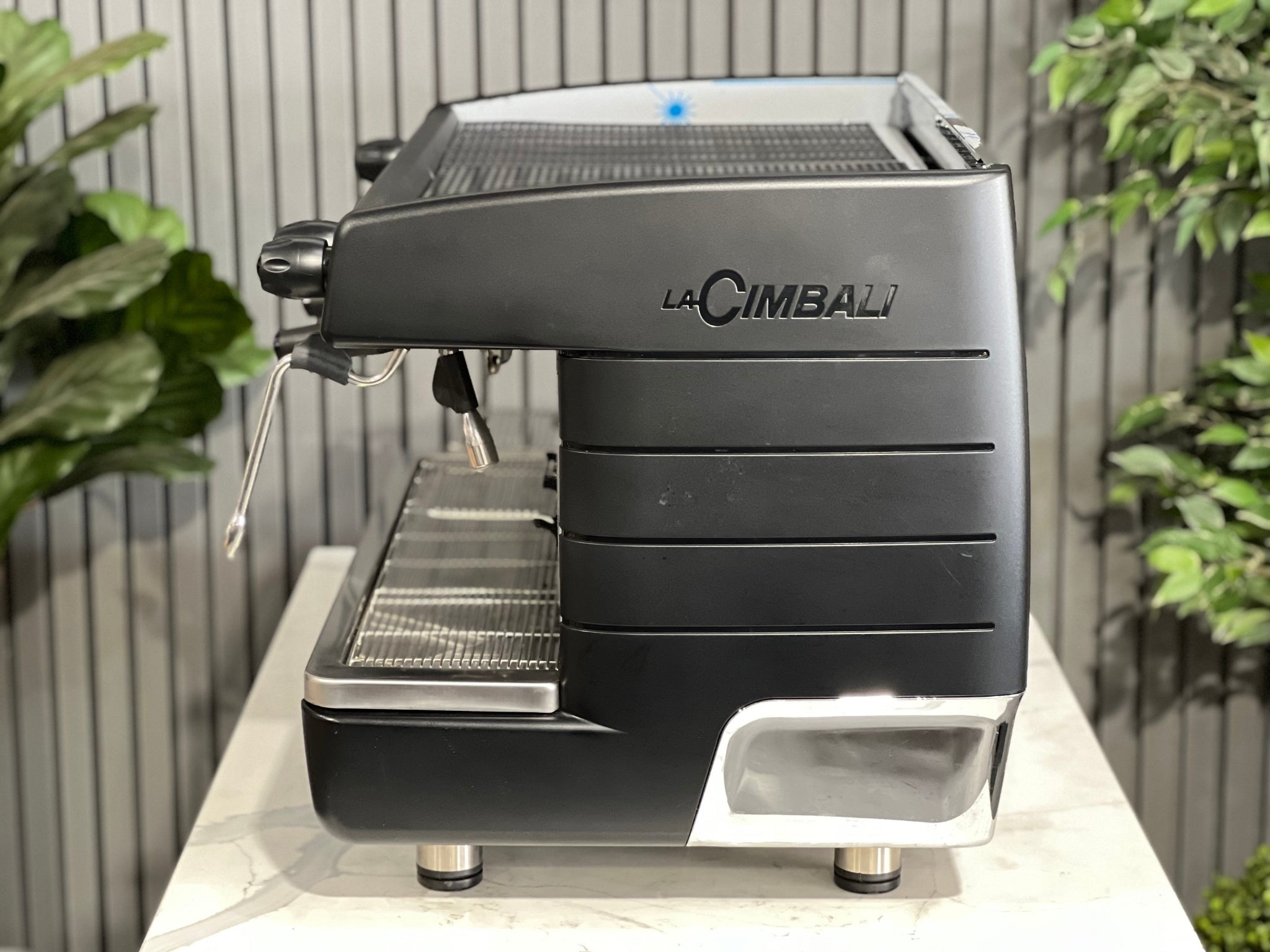 A brand new La Cimbali M23 UP 2 Group Coffee Machine with Black finishsitting on a marble bench. There is greenery in the background. The image is taken from the side of the machine.
