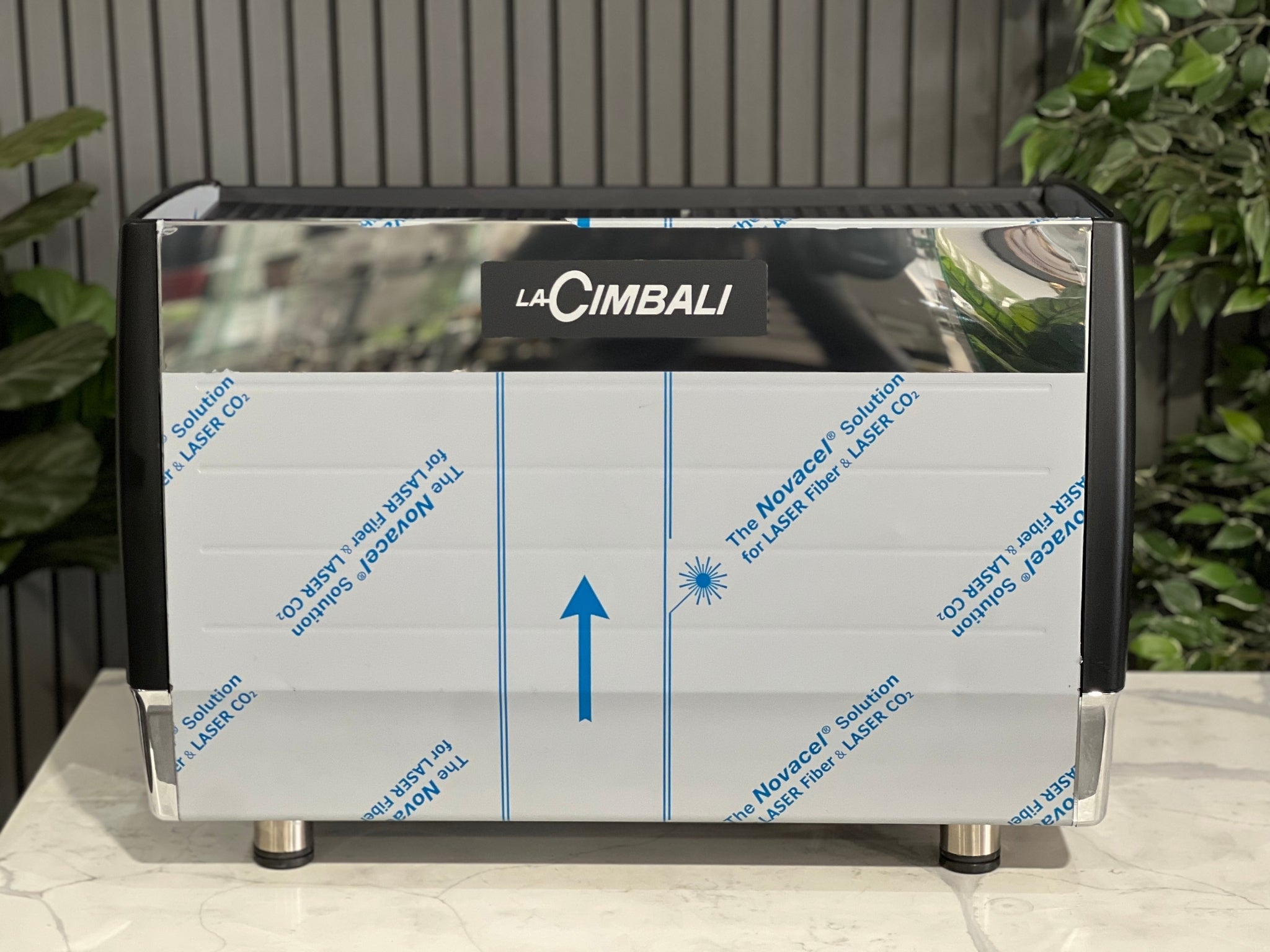 A brand new La Cimbali M23 UP 2 Group Coffee Machine with Black finish
 is sitting on a marble bench. There is greenery in the background. The image is taken from behind the machine so the back is visible. 