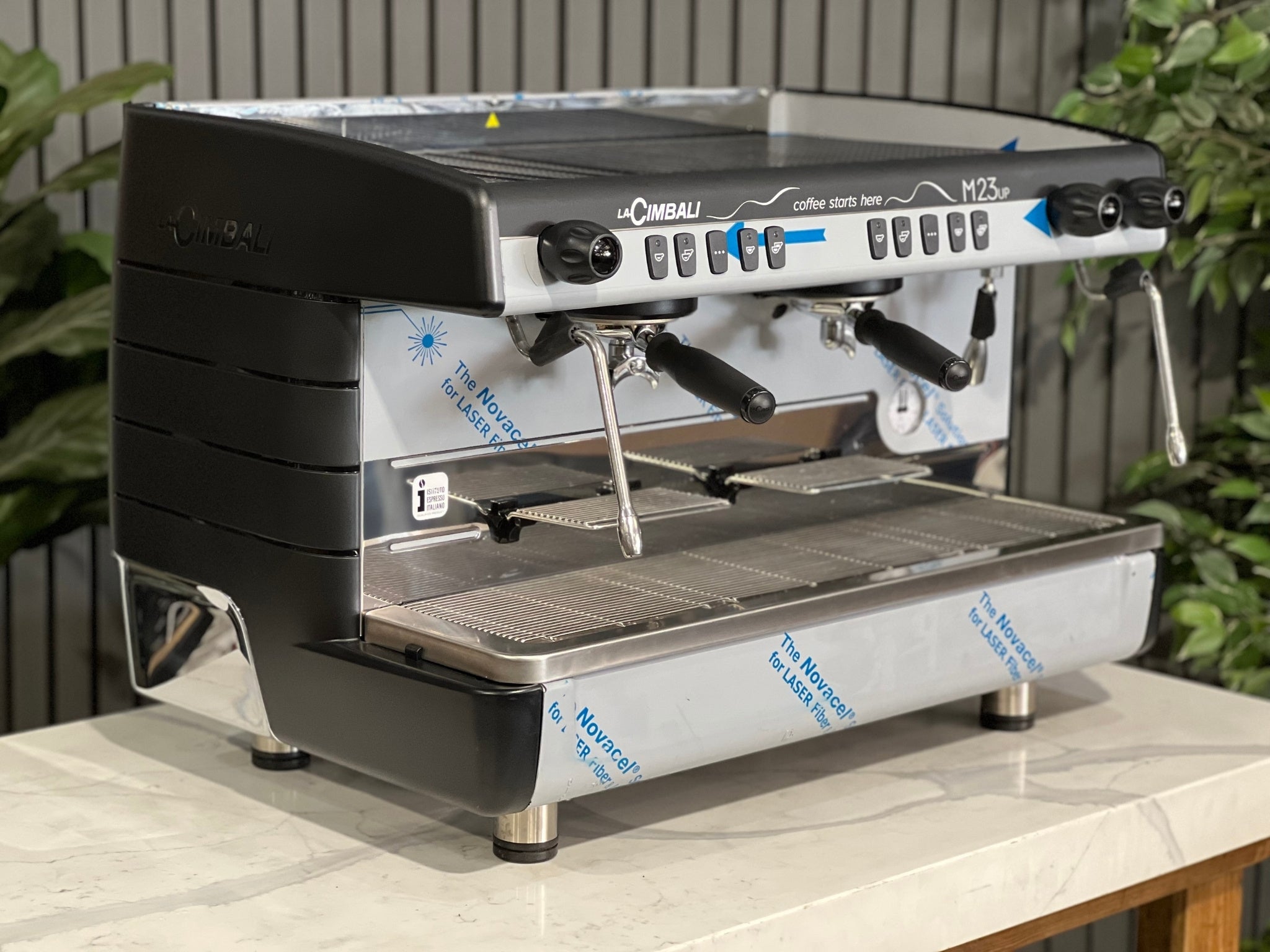 A brand new La Cimbali M23 UP 2 Group Coffee Machine with Black finish s sitting on a marble bench. There is greenery in the background. The image is taken from the front of the machine and the machine is sitting on an angle so that you can see the front of the machine and the side. 