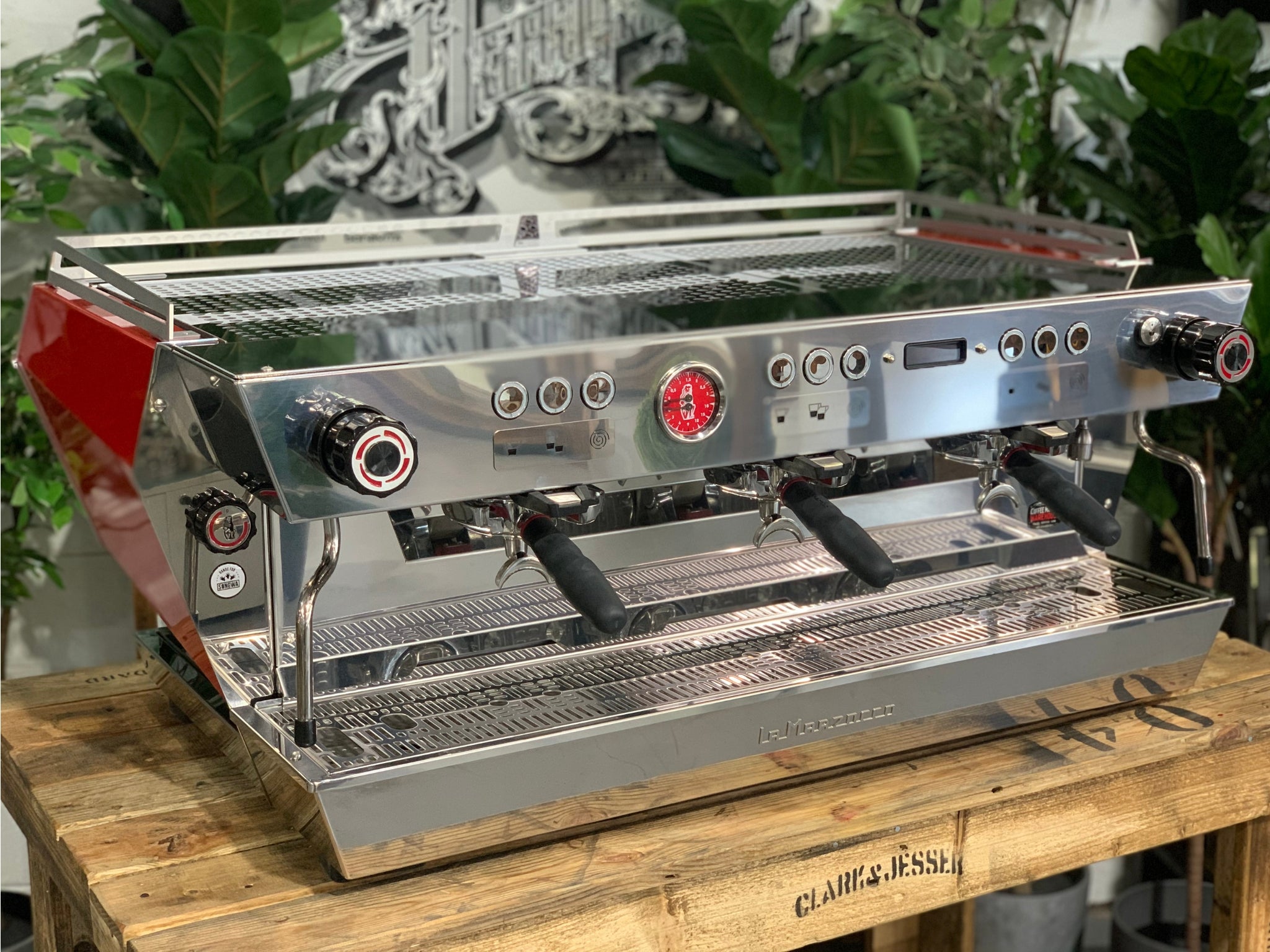 La Marzocco KB90 3 Group Red & Stainless Coffee Machine - New angled view showing three group heads, ergonomic straight-in portafilters, and stainless steel body on a wooden bench with greenery in the background, espresso coffee machine.