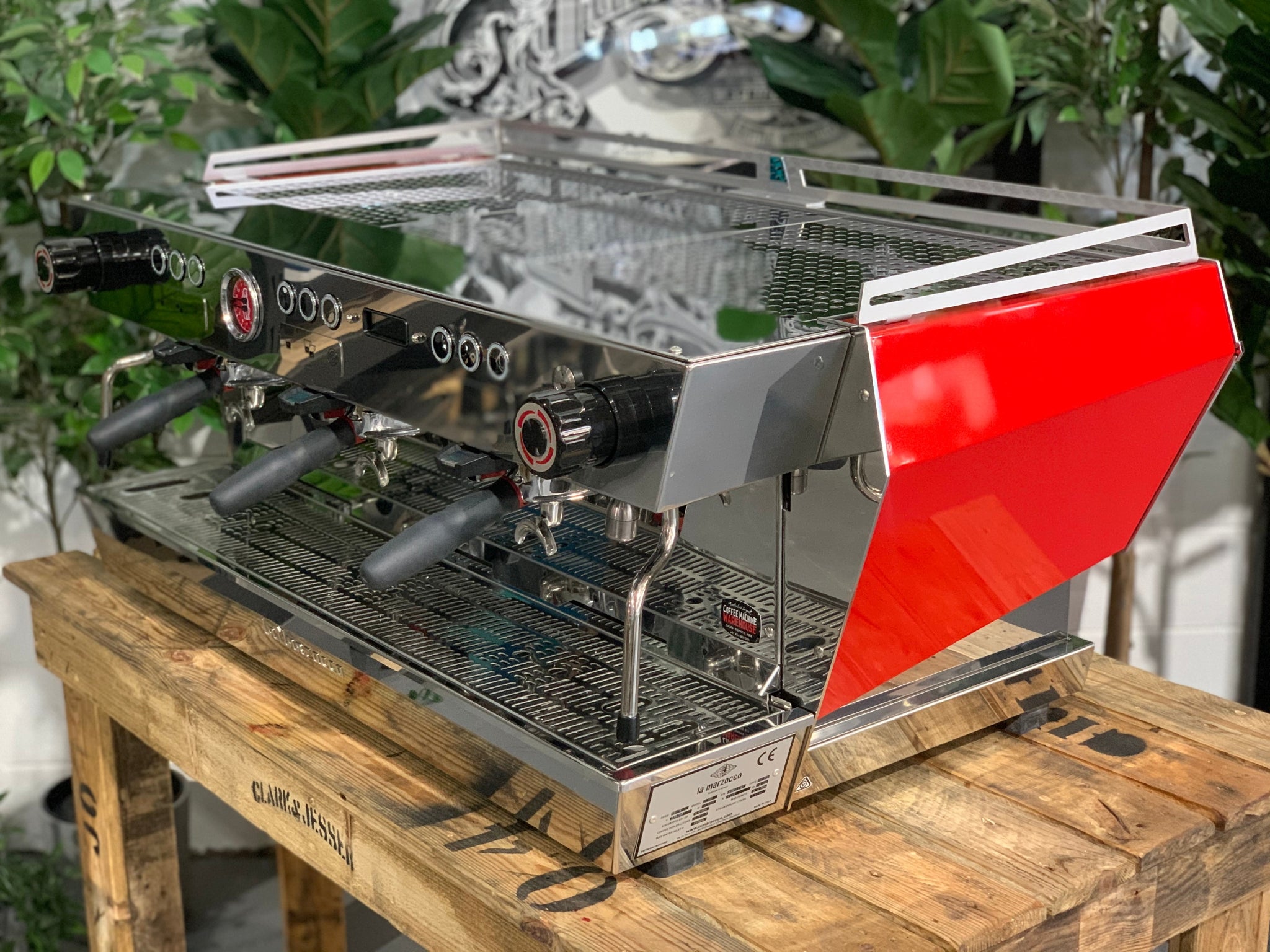 La Marzocco KB90 3 Group Red & Stainless Coffee Machine - New angled view showing three group heads, ergonomic straight-in portafilters, and stainless steel body on a wooden bench with greenery in the background, espresso coffee machine.