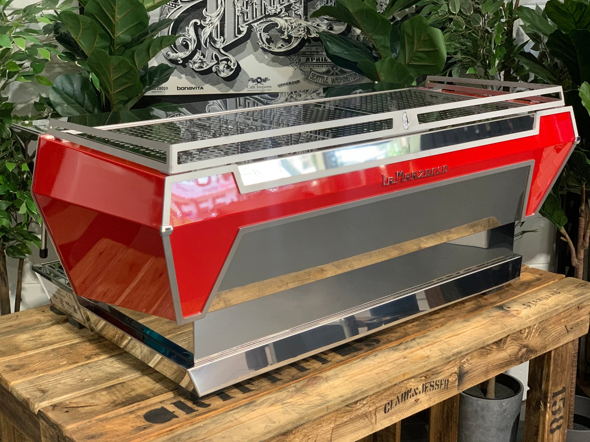 Angled rear view of La Marzocco KB90 3 Group Red & Stainless Coffee Machine - New showing side and back panels and stainless steel finish on a wooden bench with greenery, espresso machine.