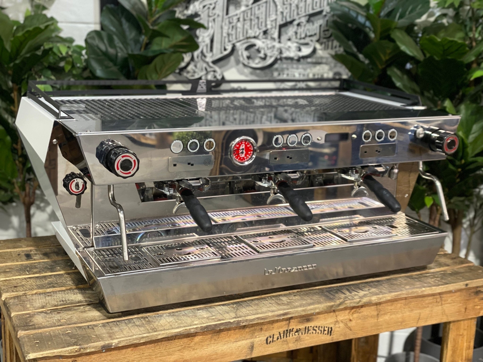 La Marzocco KB90 ABR 3 Group Coffee Machine White angled view showing the sleek white body, triple group heads, and cool touch steam wands of this high-performance commercial espresso machine, sitting on a wooden bench with greenery in the background.