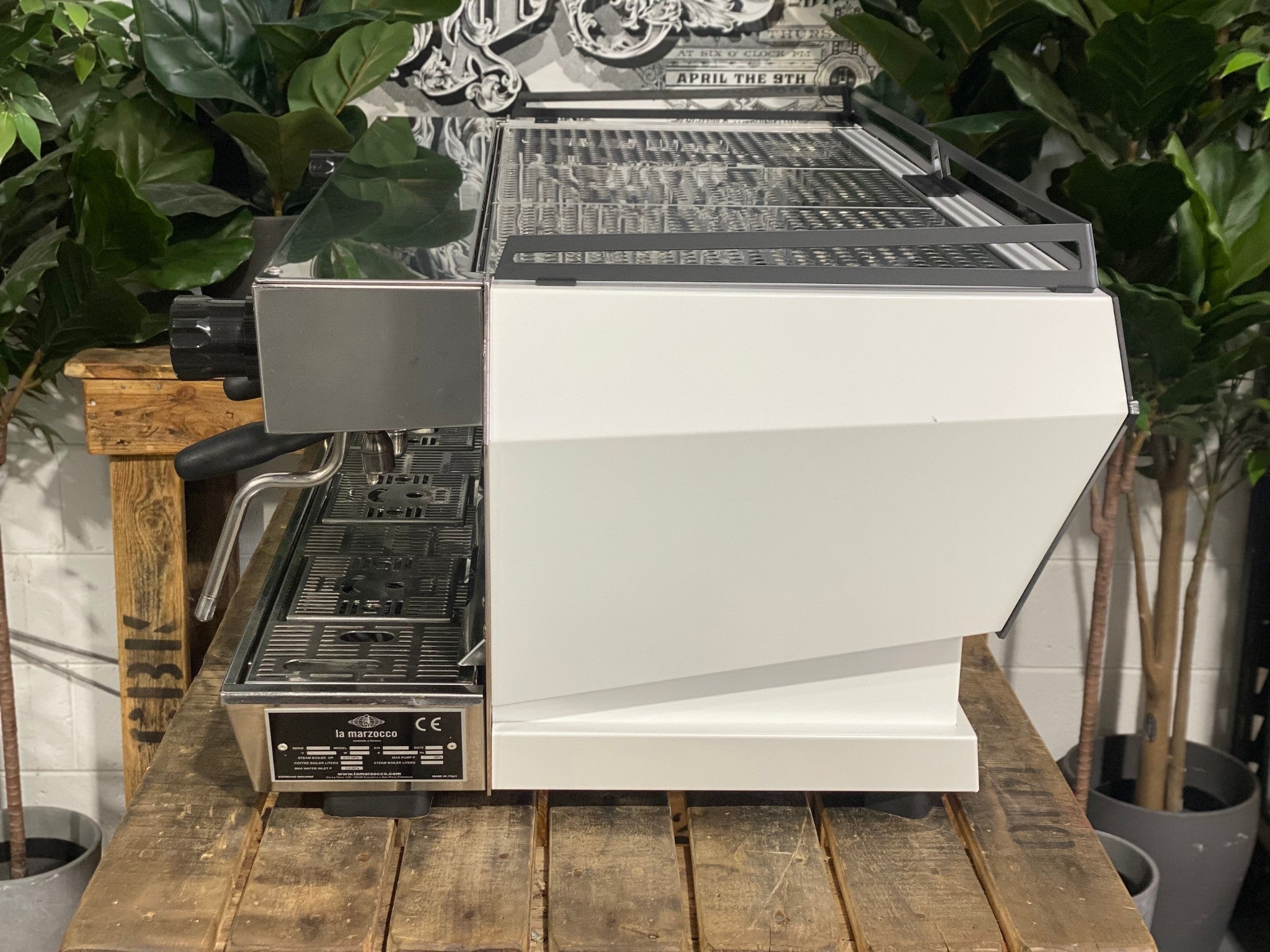 La Marzocco KB90 ABR 3 Group Coffee Machine White side view revealing the smooth side panel, cool touch steam wand, and part of the group head, designed for heavy-duty espresso performance on a wooden bench with greenery in the background.