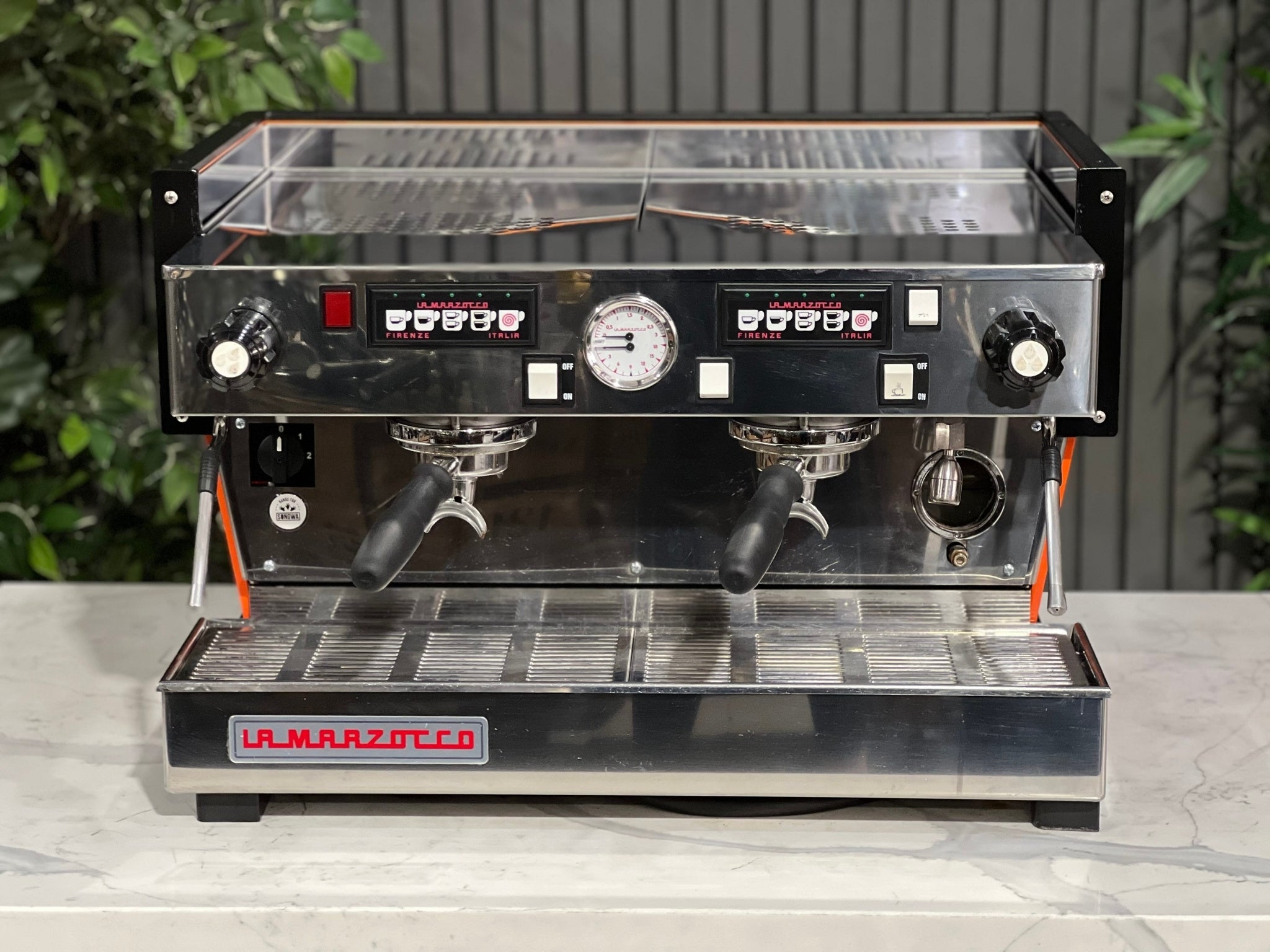 La Marzocco Linea Classic 2 Group Espresso Coffee Machine with a custom Orange finish sitting on a marble bench. There is greenery in the background. The image is taken from the front of the machine so that you can see the front of the machine, control panel, buttons and group heads.
