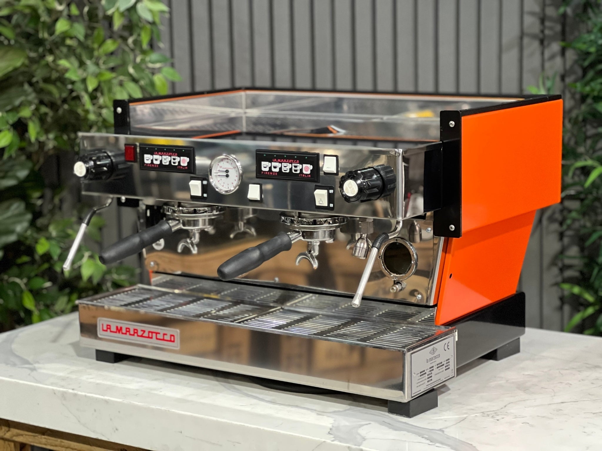 La Marzocco Linea Classic 2 Group Espresso Coffee Machine with a custom Orange finish is sitting on a marble bench. There is greenery in the background. The image is taken from the front of the machine and the machine is sitting on an angle so that you can see the front of the machine and the side. 