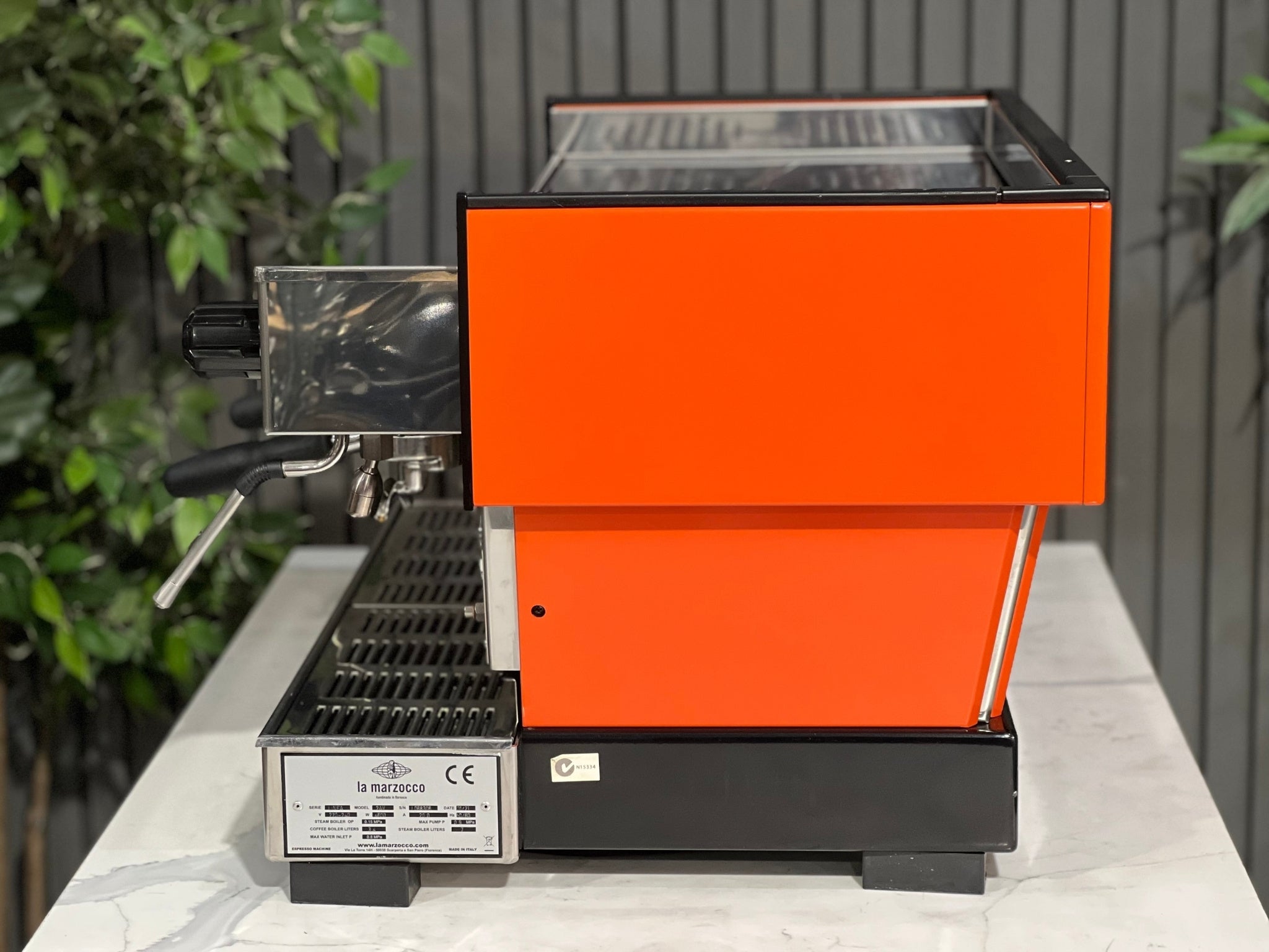 La Marzocco Linea Classic 2 Group Espresso Coffee Machine with a custom Orange finish sitting on a marble bench. There is greenery in the background. The image is taken from the side of the machine.