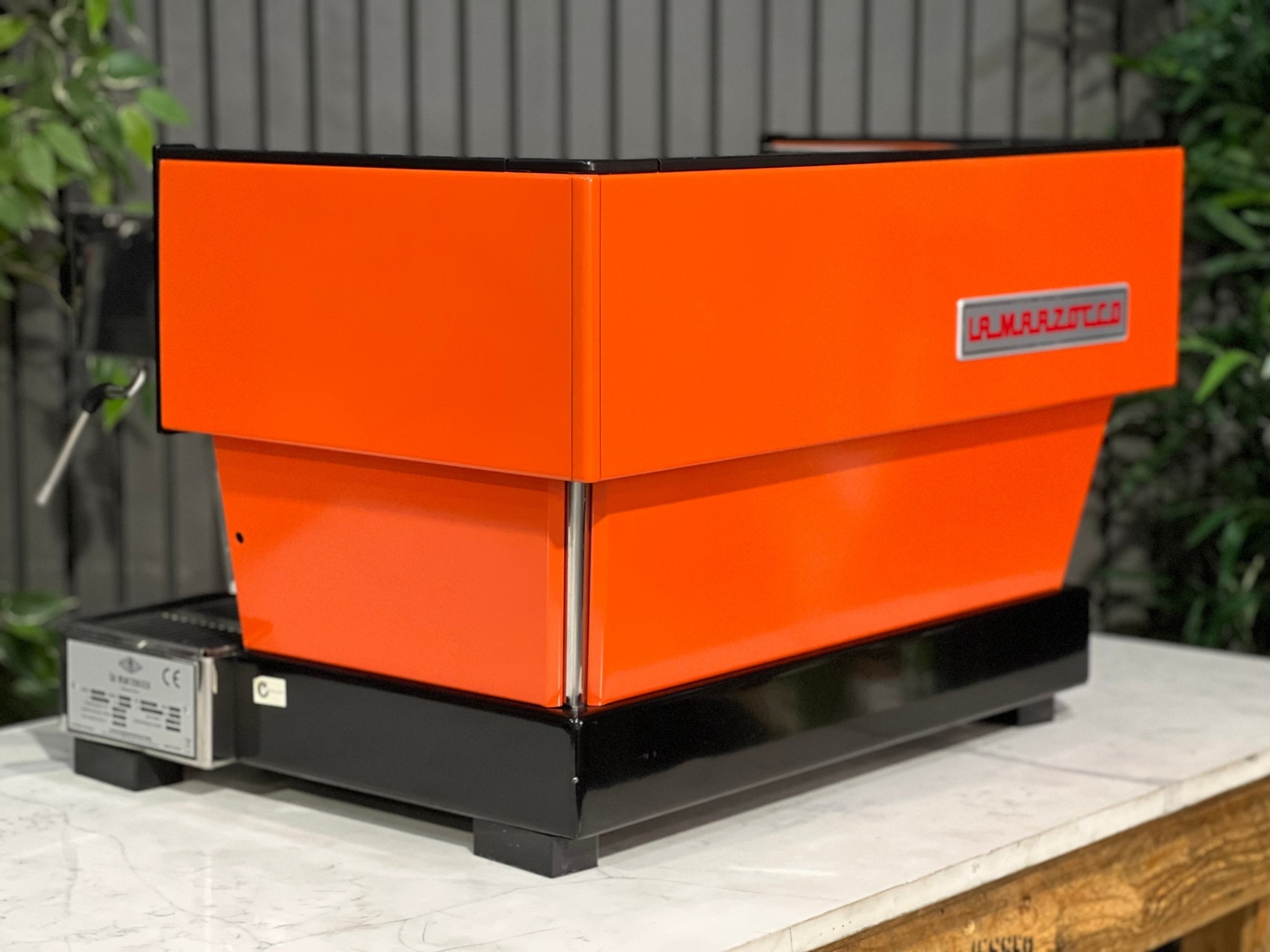 La Marzocco Linea Classic 2 Group Espresso Coffee Machine with a custom Orange finish sitting on a marble bench. There is greenery in the background. The image is taken from behind the machine and the machine is sitting on an angle so that you can see the back of the machine and the side.
