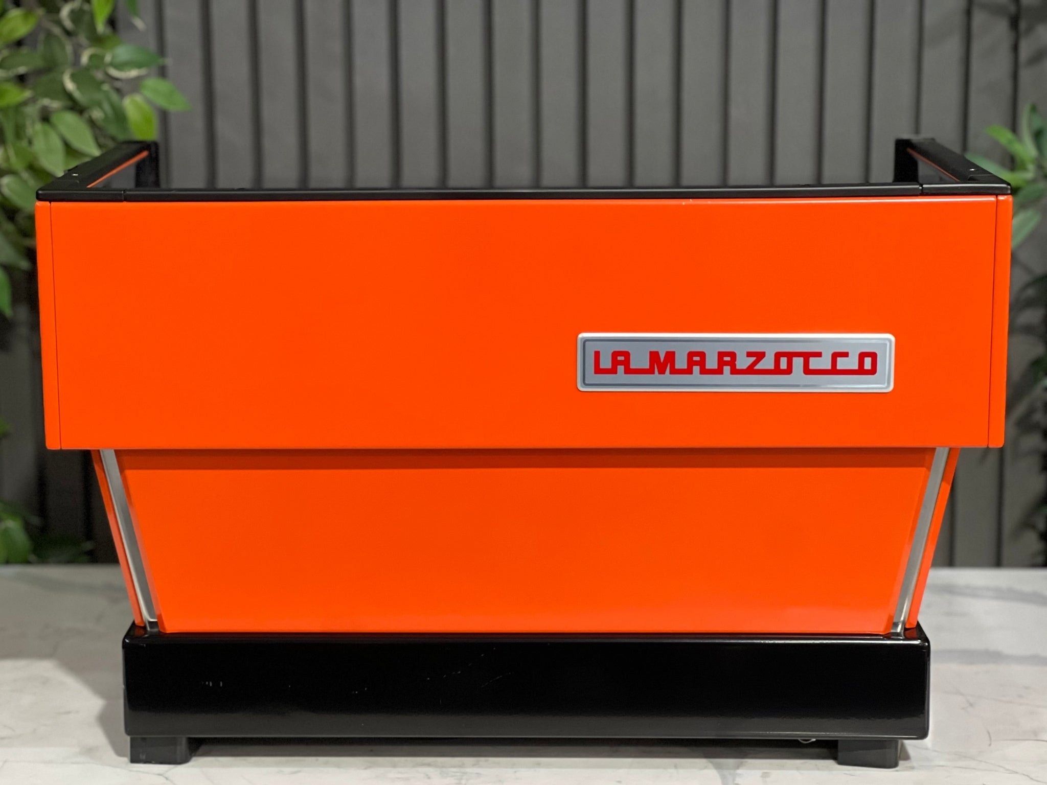 La Marzocco Linea Classic 2 Group Espresso Coffee Machine with a custom Orange finish is sitting on a marble bench. There is greenery in the background. The image is taken from behind the machine so the back is visible. The back of the machine is a orange with the red la marzocco logo on a silver background. The logo is situated on the right hand side of the machine.
