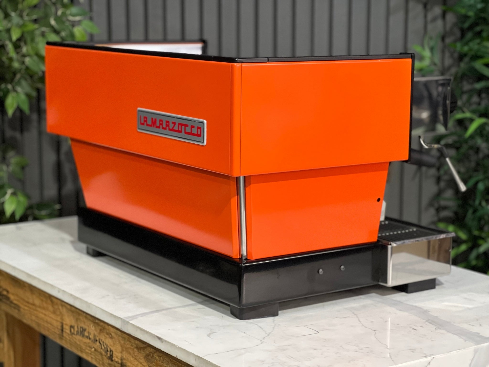 La Marzocco Linea Classic 2 Group Espresso Coffee Machine with a custom Orange finish sitting on a marble bench. There is greenery in the background. The image is taken from behind the machine and the machine is sitting on an angle so that you can see the back of the machine and the side.
