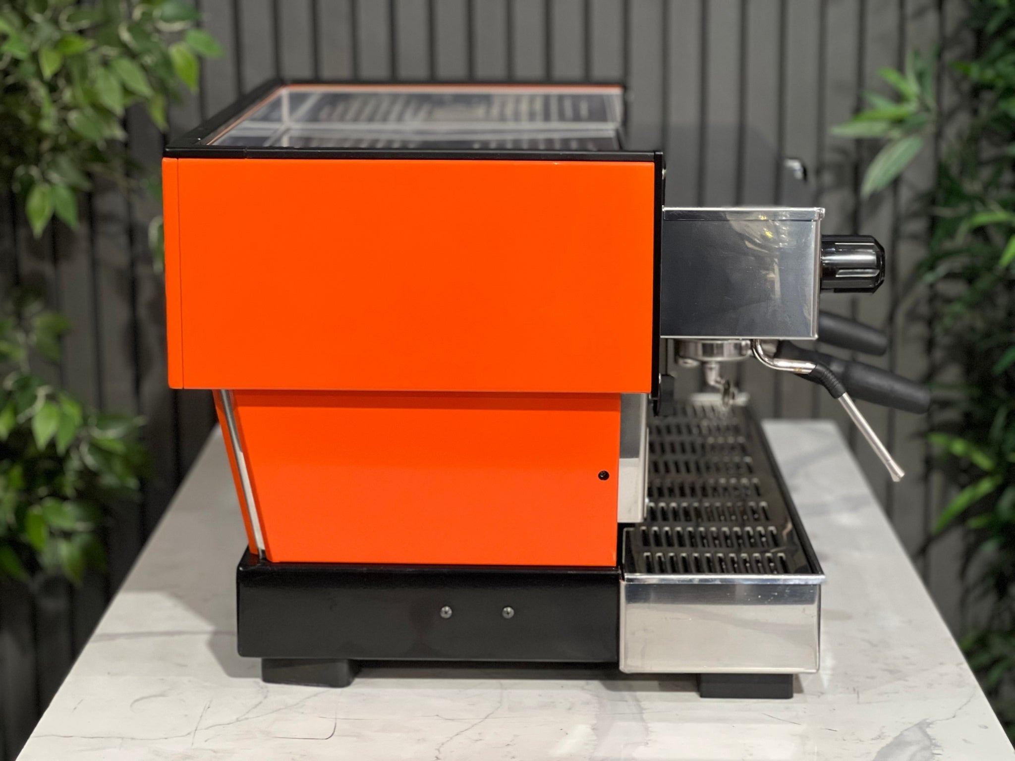 La Marzocco Linea Classic 2 Group Espresso Coffee Machine with a custom Orange finish sitting on a marble bench. There is greenery in the background. The image is taken from the side of the machine.
