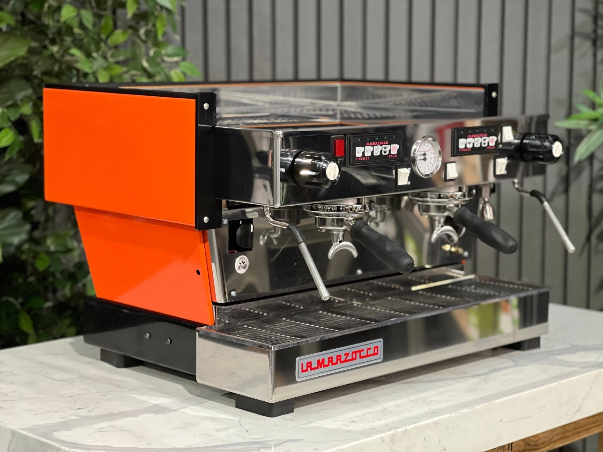 La Marzocco Linea Classic 2 Group Espresso Coffee Machine with a custom Orange finish  is sitting on a marble bench. There is greenery in the background. The image is taken from the front of the machine and the machine is sitting on an angle so that you can see the front of the machine and the side. 