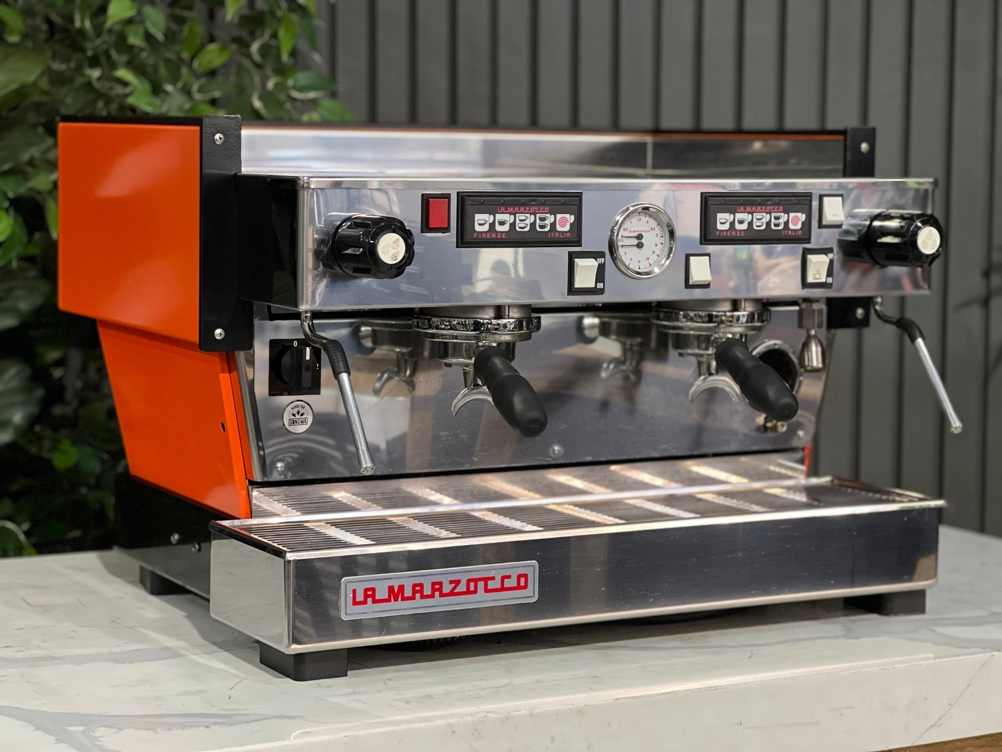 La Marzocco Linea Classic 2 Group Espresso Coffee Machine with a custom Orange finish  is sitting on a marble bench. There is greenery in the background. The image is taken from the front of the machine and the machine is sitting on an angle so that you can see the front of the machine and the side. 