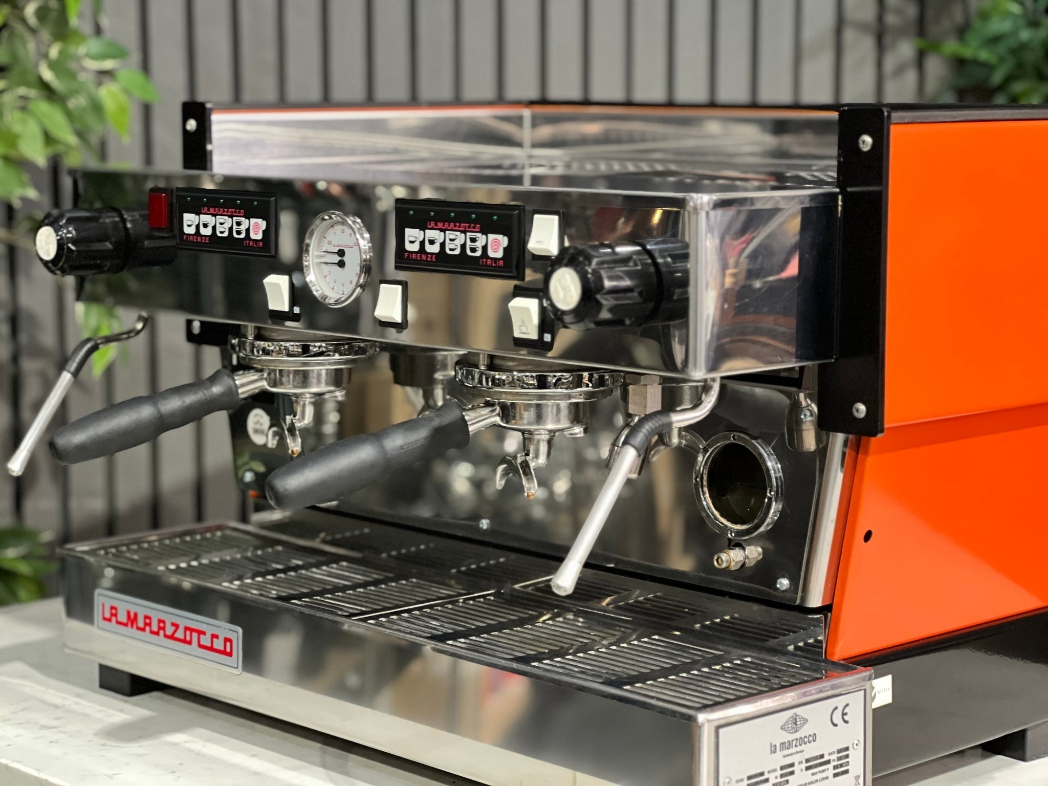 La Marzocco Linea Classic 2 Group Espresso Coffee Machine with a custom Orange finish s sitting on a marble bench. There is greenery in the background. The image is a close up image taken from the front of the machine and the machine is sitting on an angle so that you can see the front of the machine, the controls including buttons, group handles and logo.
