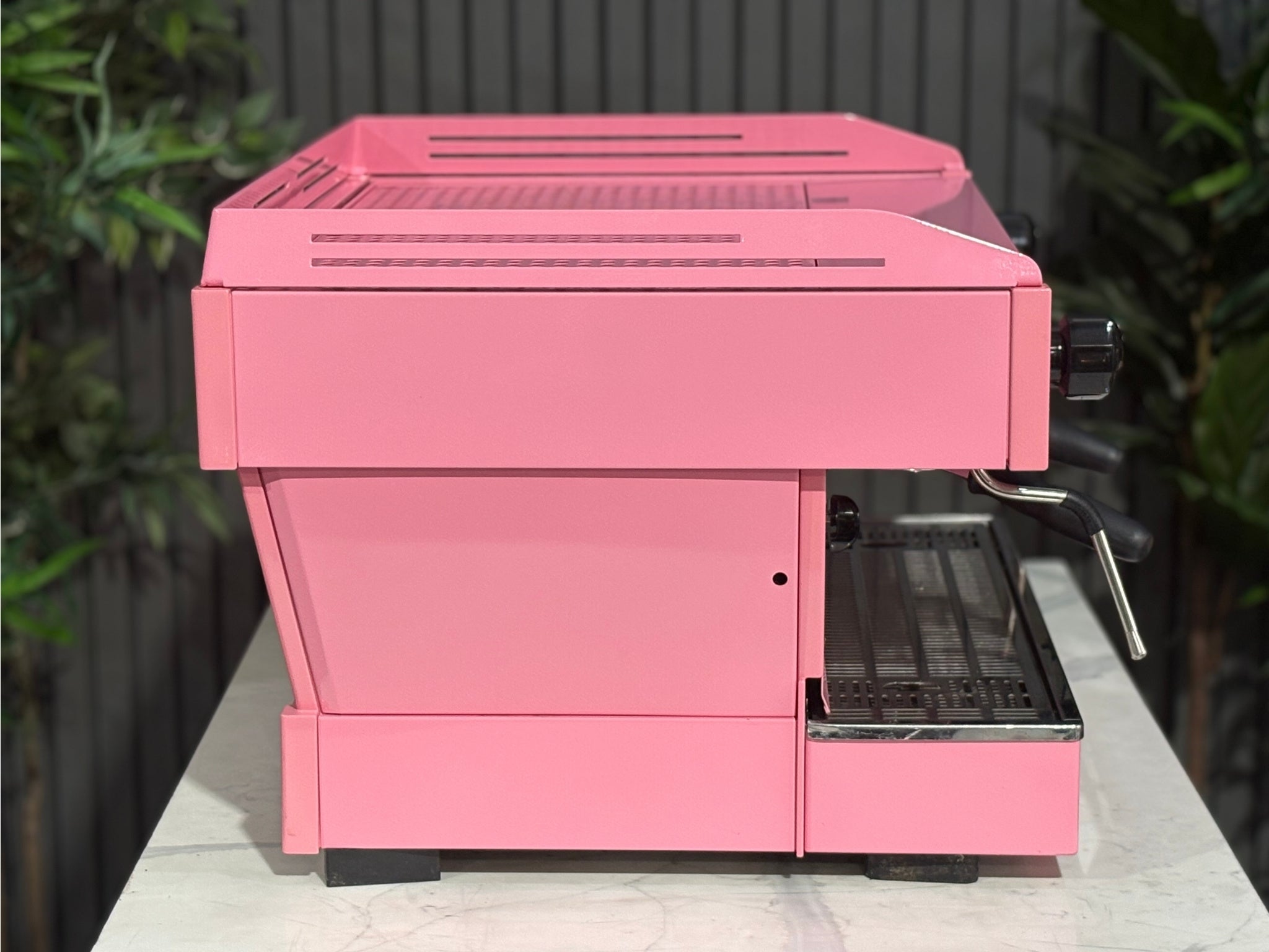 La Marzocco Linea PB 2 Group Coffee Machine Dark Pink side view revealing one group head, the dual boiler structure, and part of the steam wand, all in a vibrant pink finish on a marble bench with greenery.