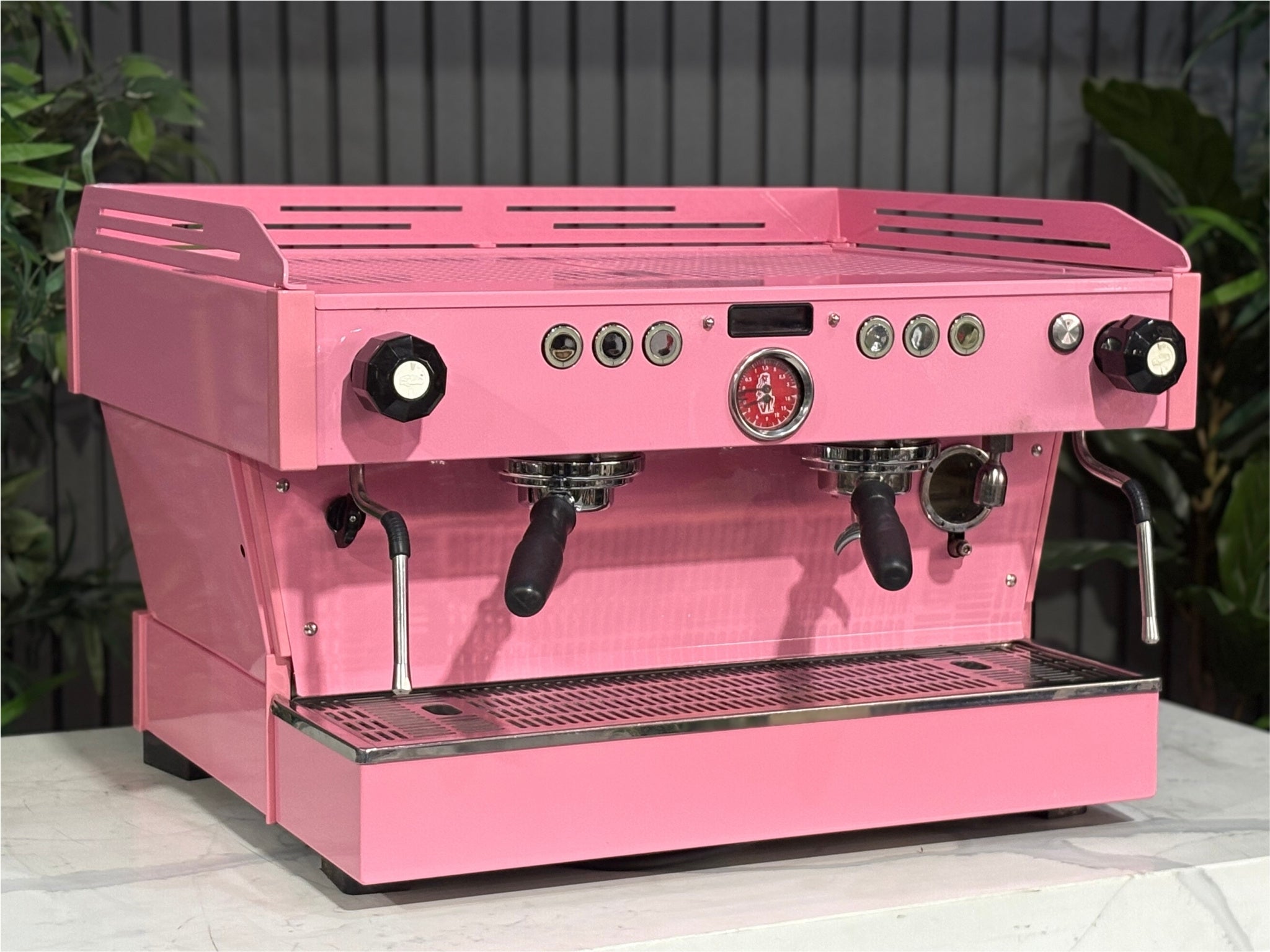 La Marzocco Linea PB 2 Group Coffee Machine Dark Pink angled view showcasing the dual group heads, LED display, and striking custom pink body of this commercial espresso coffee machine, positioned on a marble bench with greenery in the background.