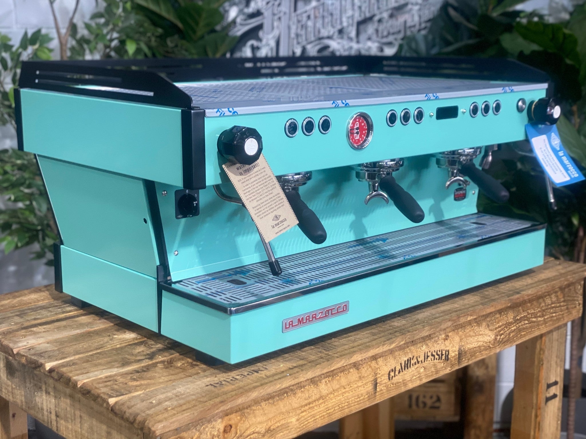 La Marzocco Linea PB 3 Group Coffee Machine Aqua - New espresso machine shown at an angled view, displaying the front and one side of the aqua body, dual steam wands, group heads, cup tray, and digital display on a wooden bench with greenery in the background.