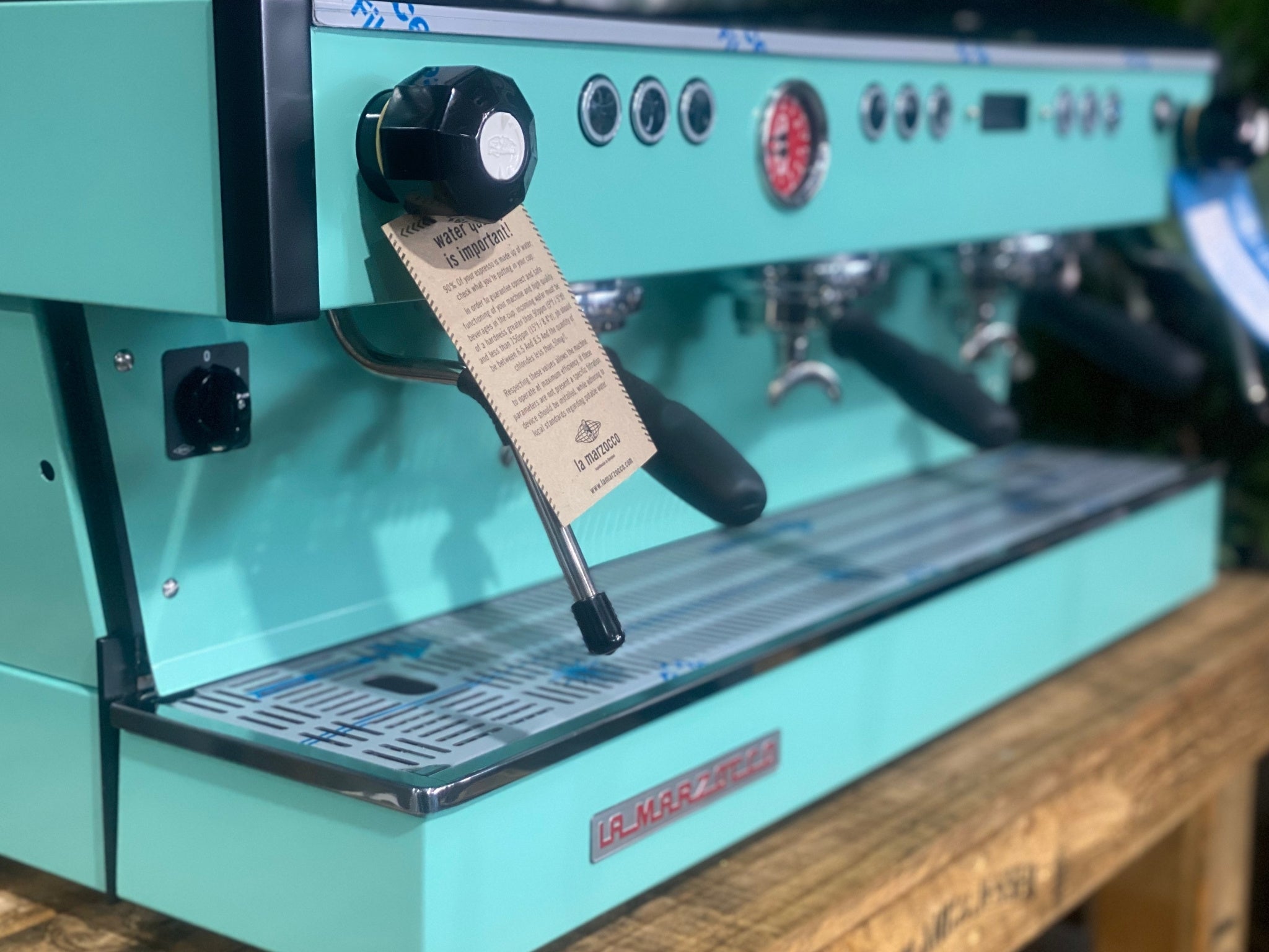 La Marzocco Linea PB 3 Group Coffee Machine Aqua - New espresso machine close-up at an angled view, showing the group heads, steam wands, cup tray, and digital display on the aqua body, set on a wooden bench with greenery in the background.