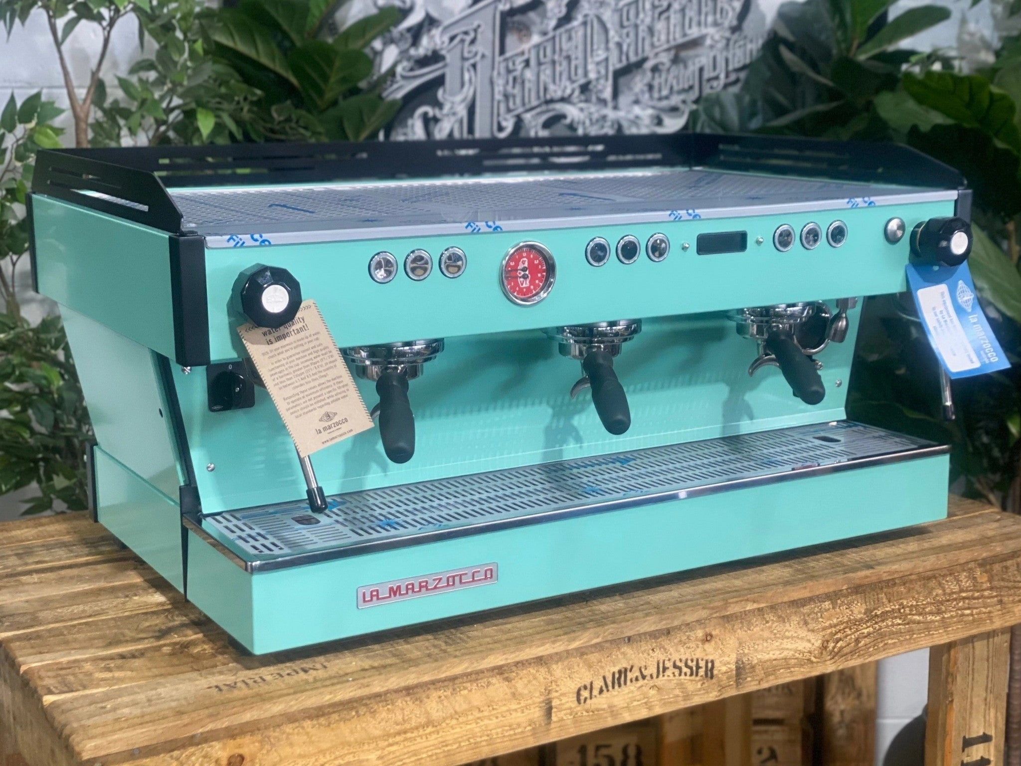 La Marzocco Linea PB 3 Group Coffee Machine Aqua - New espresso machine shown at an angled view, displaying the front and one side of the aqua body, dual steam wands, group heads, cup tray, and digital display on a wooden bench with greenery in the background.