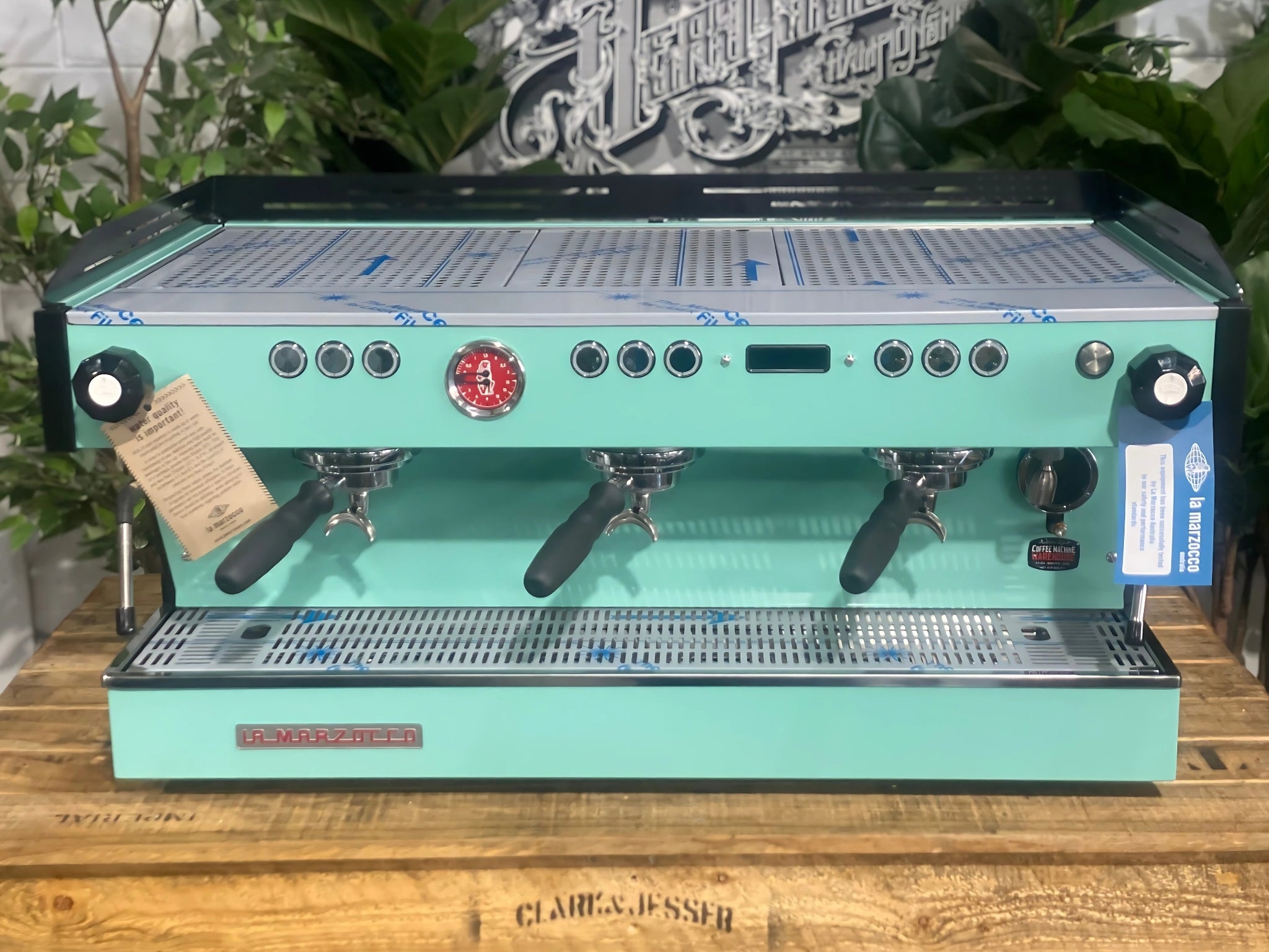 La Marzocco Linea PB 3 Group Coffee Machine Aqua - New espresso coffee machine photographed directly from the front, showing all three group heads, aqua body, cup tray, and digital display on a wooden bench with greenery behind.