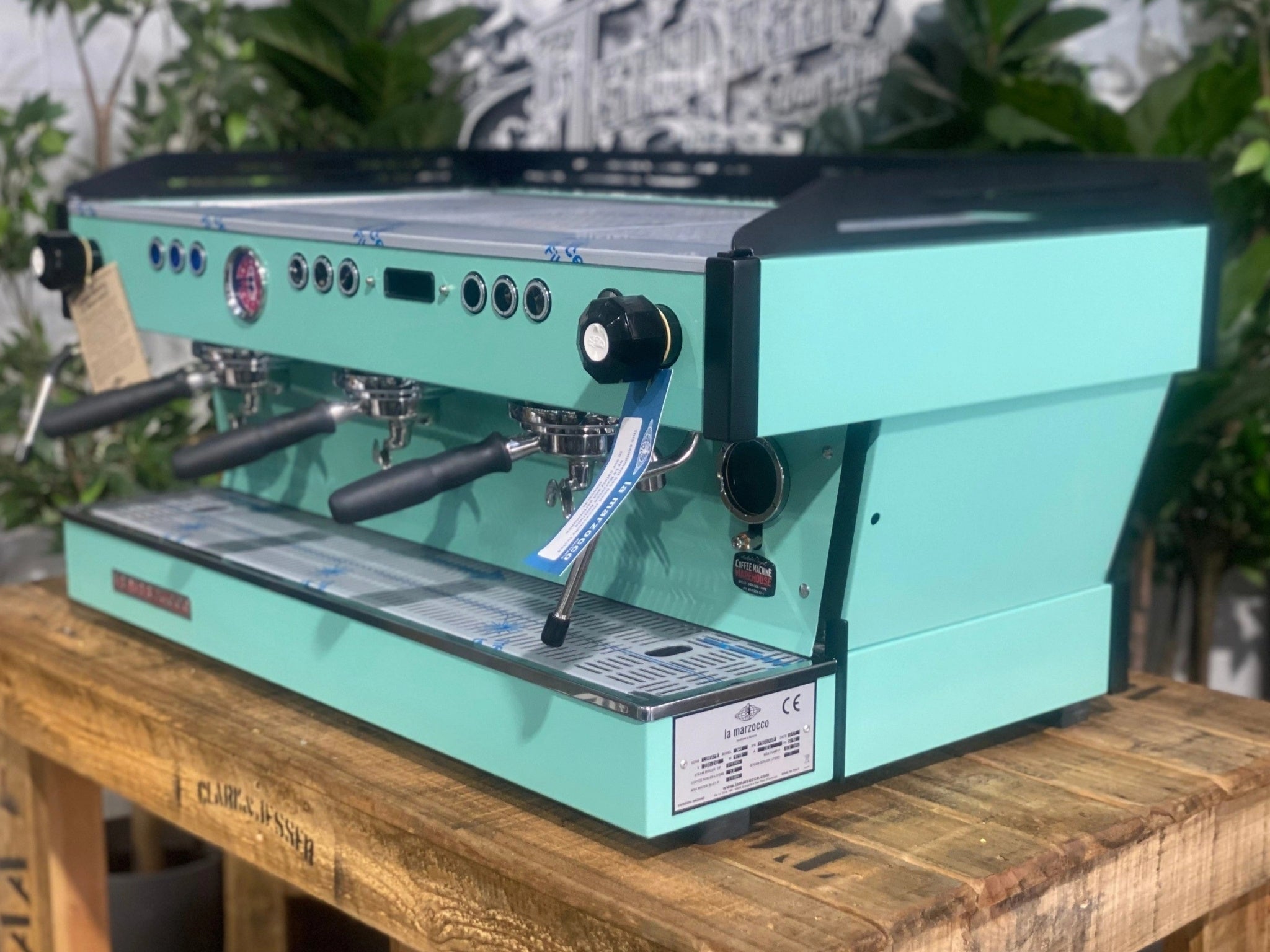 La Marzocco Linea PB 3 Group Coffee Machine Aqua - New espresso machine shown at an angled view, displaying the front and one side of the aqua body, dual steam wands, group heads, cup tray, and digital display on a wooden bench with greenery in the background.