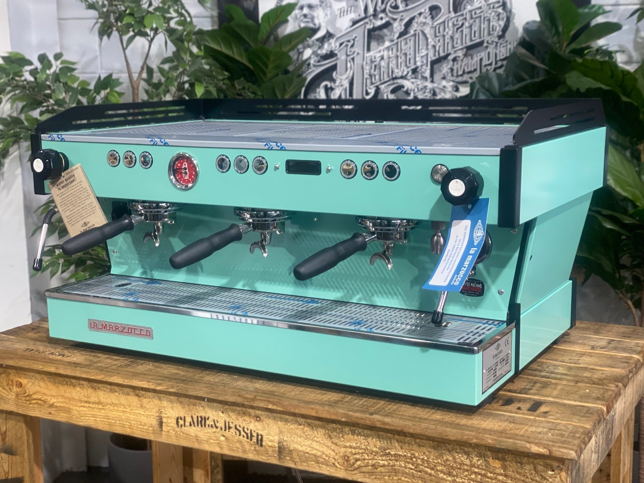 La Marzocco Linea PB 3 Group Coffee Machine Aqua - New espresso machine shown at an angled view, displaying the front and one side of the aqua body, dual steam wands, group heads, cup tray, and digital display on a wooden bench with greenery in the background.
