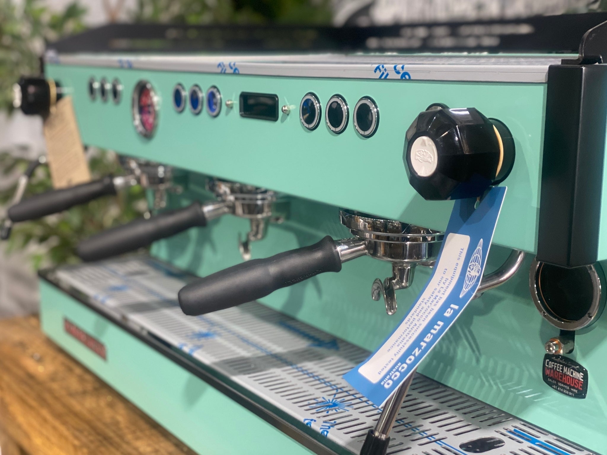 La Marzocco Linea PB 3 Group Coffee Machine Aqua - New espresso machine close-up at an angled view, showing the group heads, steam wands, cup tray, and digital display on the aqua body, set on a wooden bench with greenery in the background.