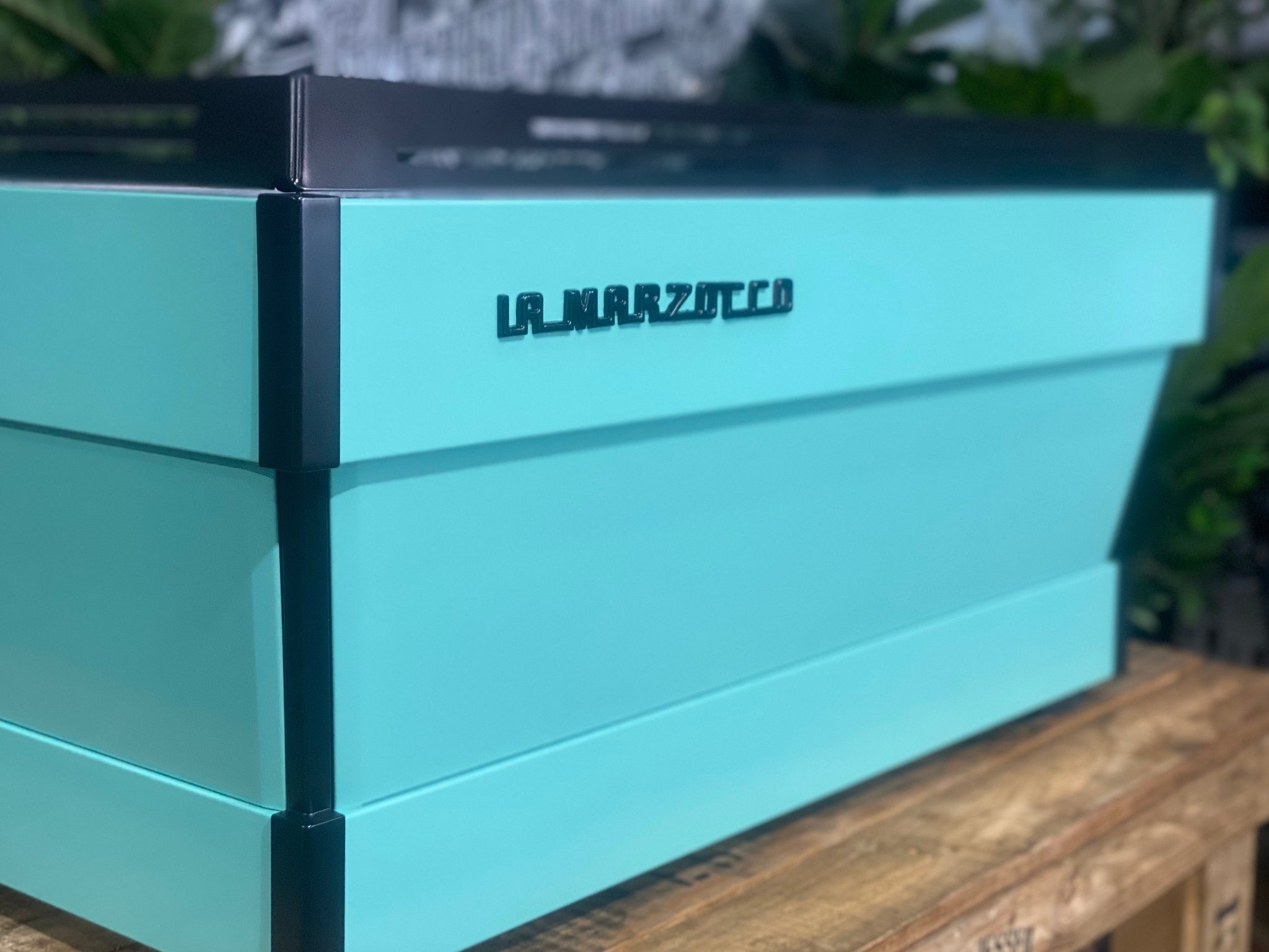 La Marzocco Linea PB 3 Group Coffee Machine Aqua - New espresso coffee machine shown at an angled rear view, highlighting the back and side panels, aqua body, dual steam wands, and cup tray on a wooden bench with greenery.