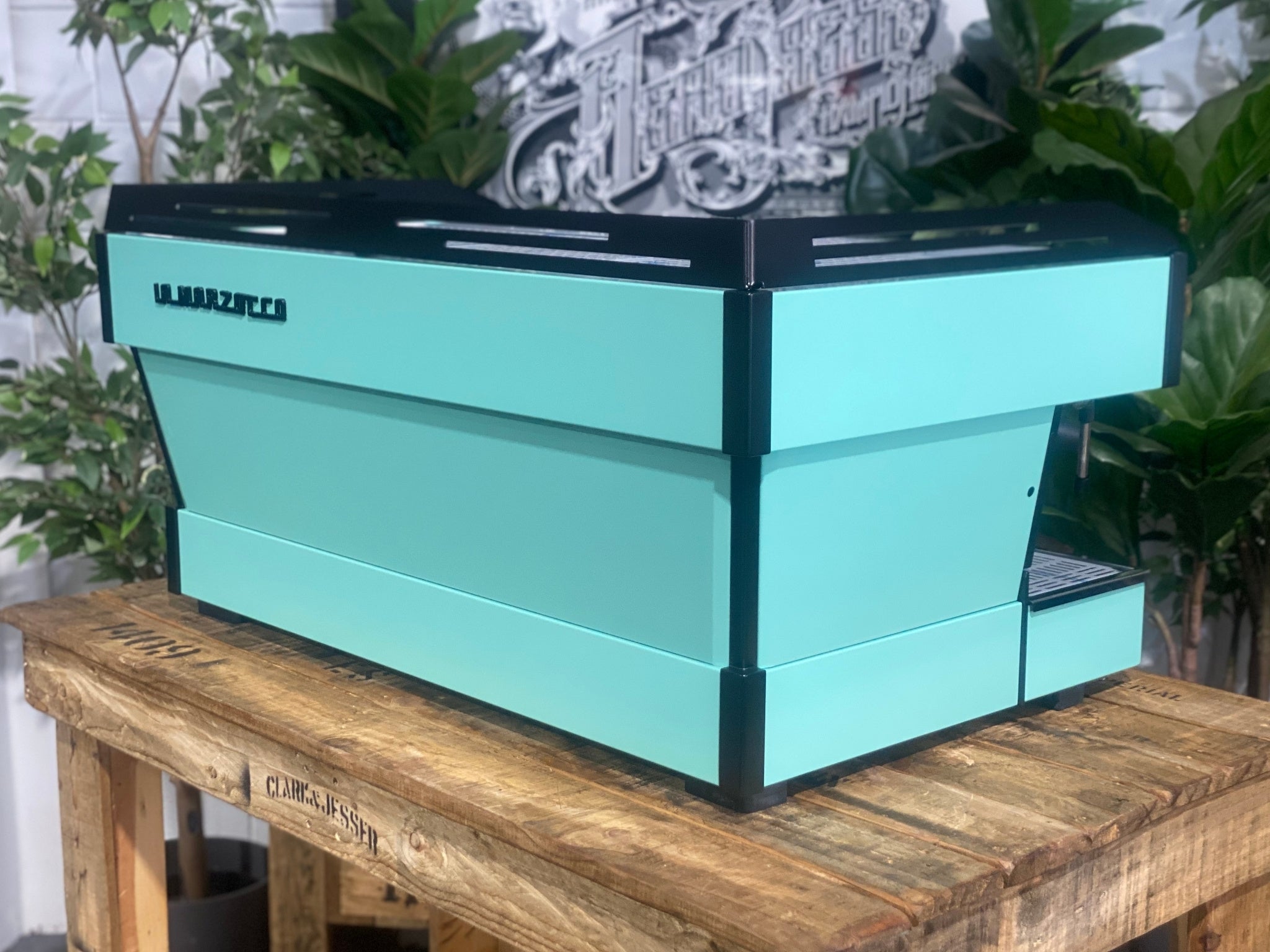 La Marzocco Linea PB 3 Group Coffee Machine Aqua - New espresso coffee machine shown at an angled rear view, highlighting the back and side panels, aqua body, dual steam wands, and cup tray on a wooden bench with greenery.